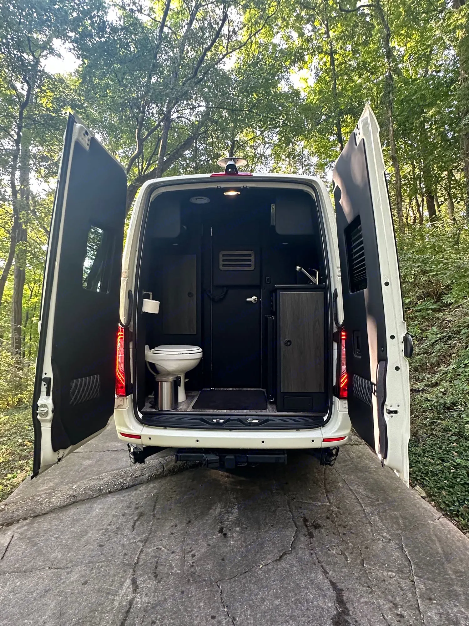 RV rental interior: Back of a white camper van with open doors revealing a compact toilet and sink area,. Mercedes-Benz Sprinter 419 LWB High Roof 2024