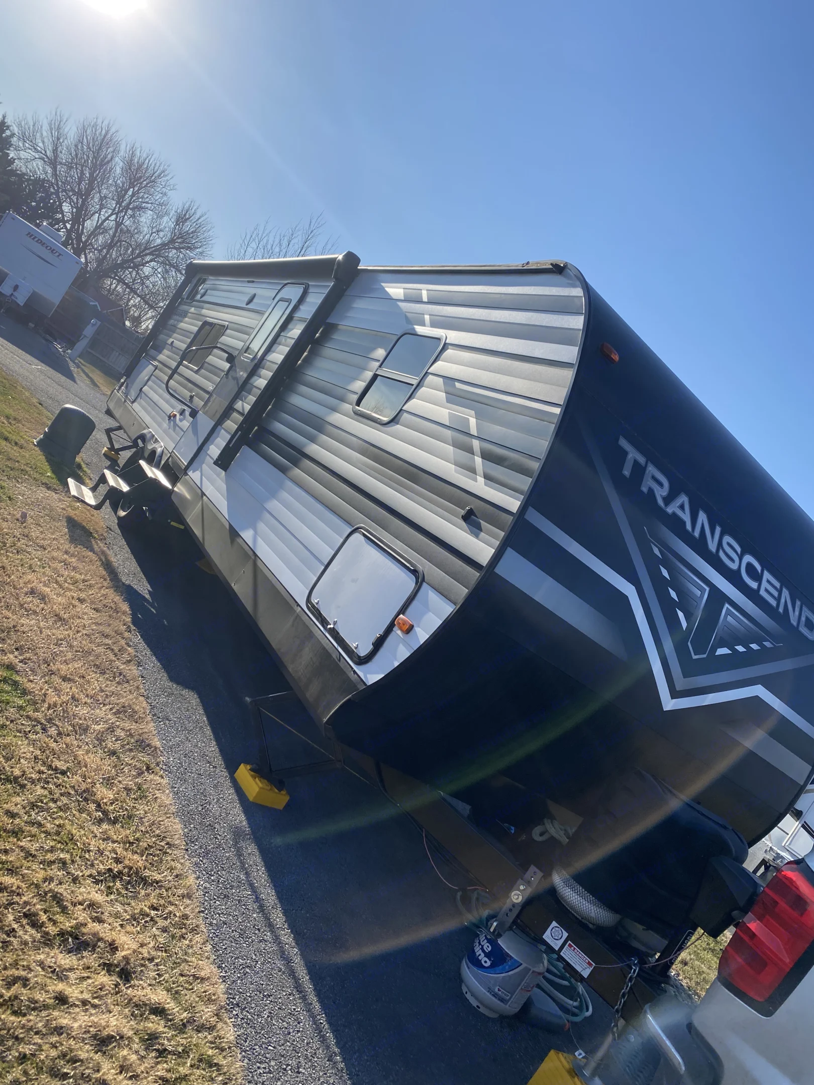 A large grey and black Transcend travel trailer with propane tank, ideal for an rv rental, parked on gravel under a bright sun.. Grand Design Transcend Xplor 2021