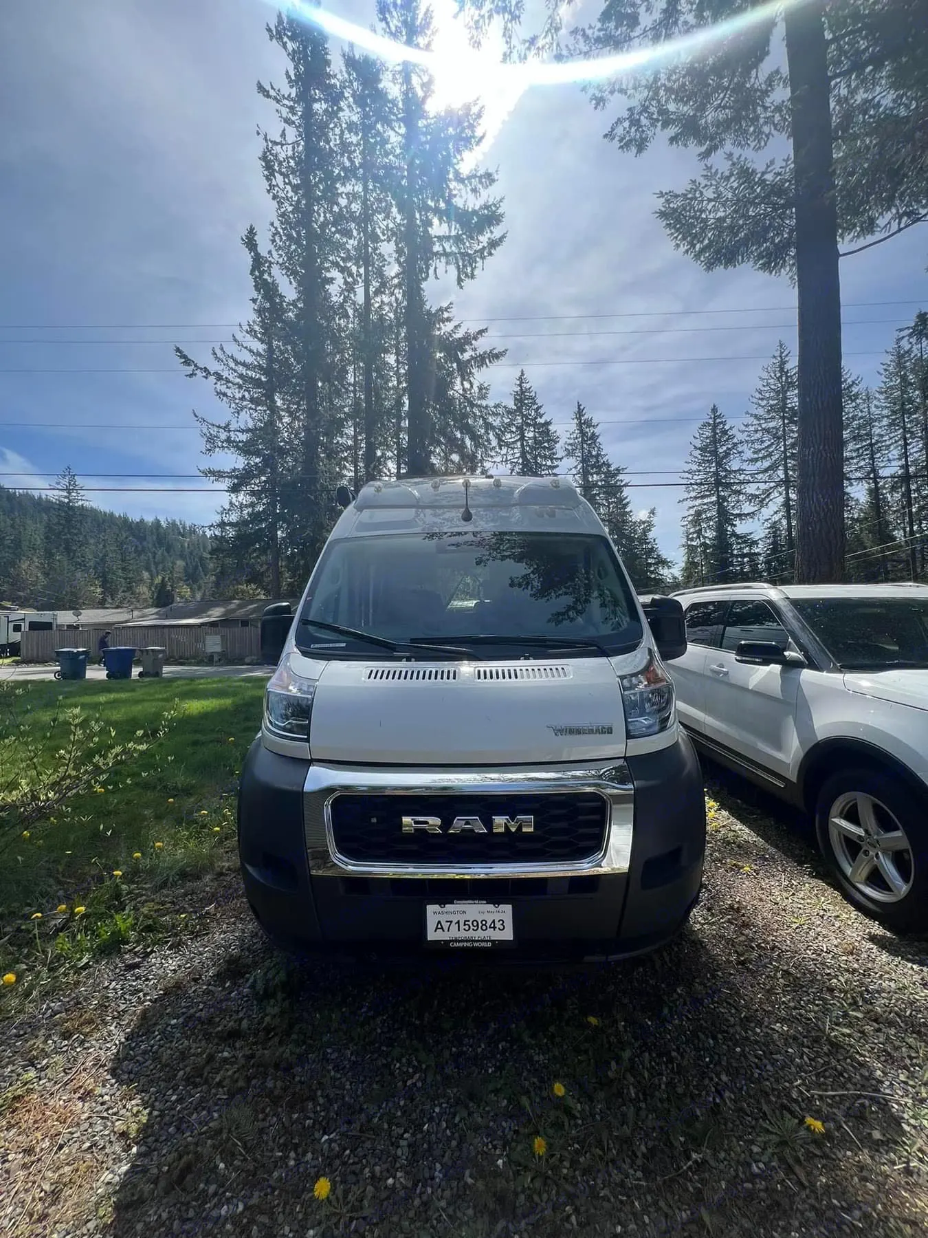 Front view of a white Ram ProMaster RV rental parked on a sunny gravel lot, surrounded by tall evergreen trees.. Winnebago Solis 2023