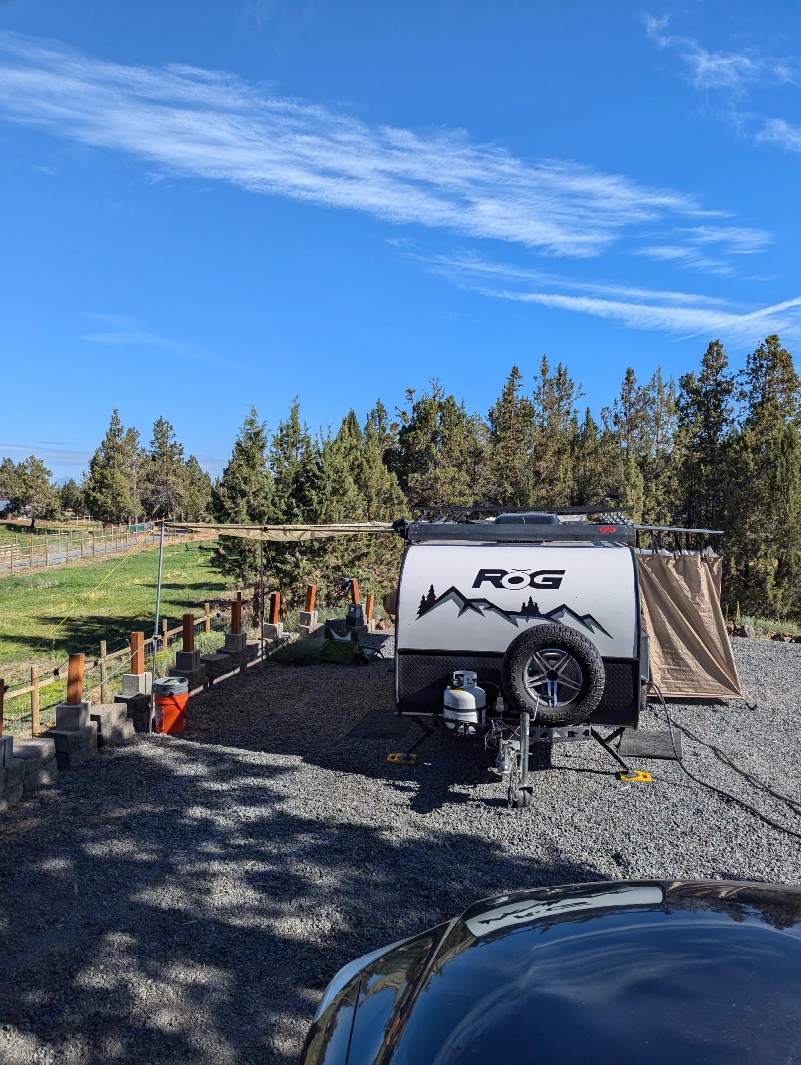 A white compact travel trailer, great for an **RV rental**, is parked on a sunny gravel campsite with an awning extended, surrounded by trees.. Encore RV ROG 2022