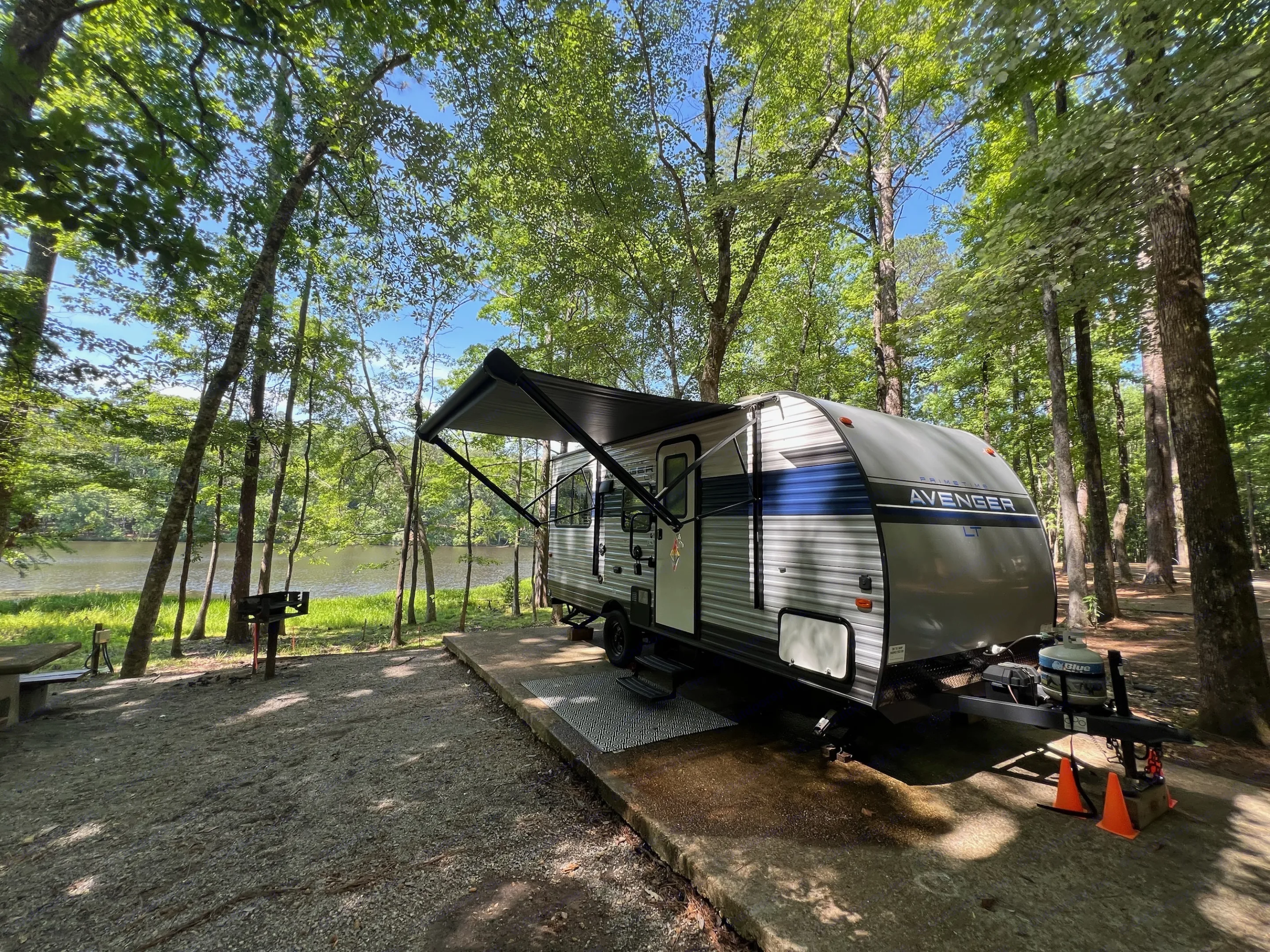 A grey RV rental with awning extended, parked on a concrete pad by a calm lake at a tree-filled campsite.. Avenger LT 2022
