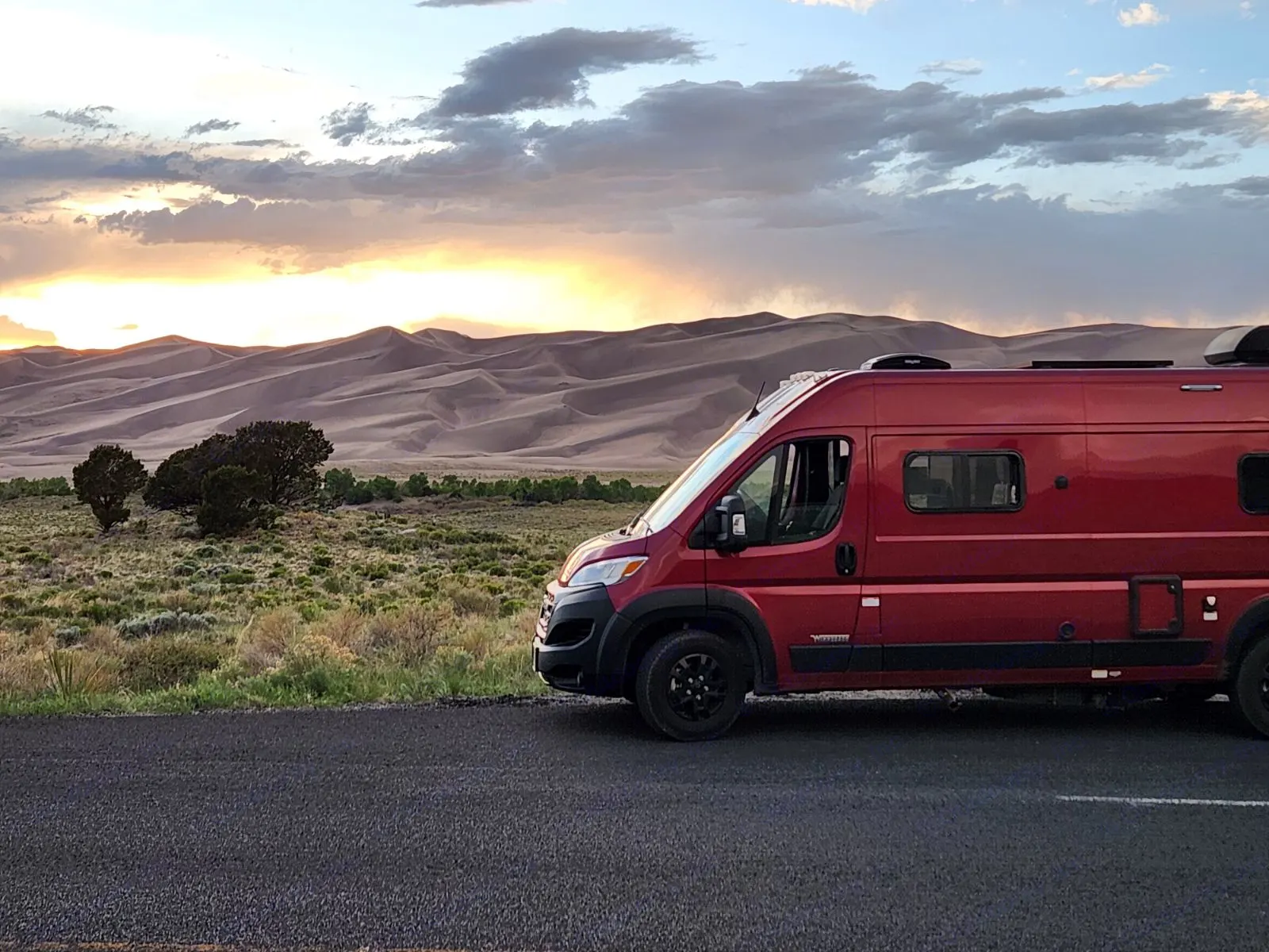 A red RV rental parked on a road, facing golden sand dunes under a colorful sunset sky. Perfect for adventure travel.. Winnebago Solis 2023