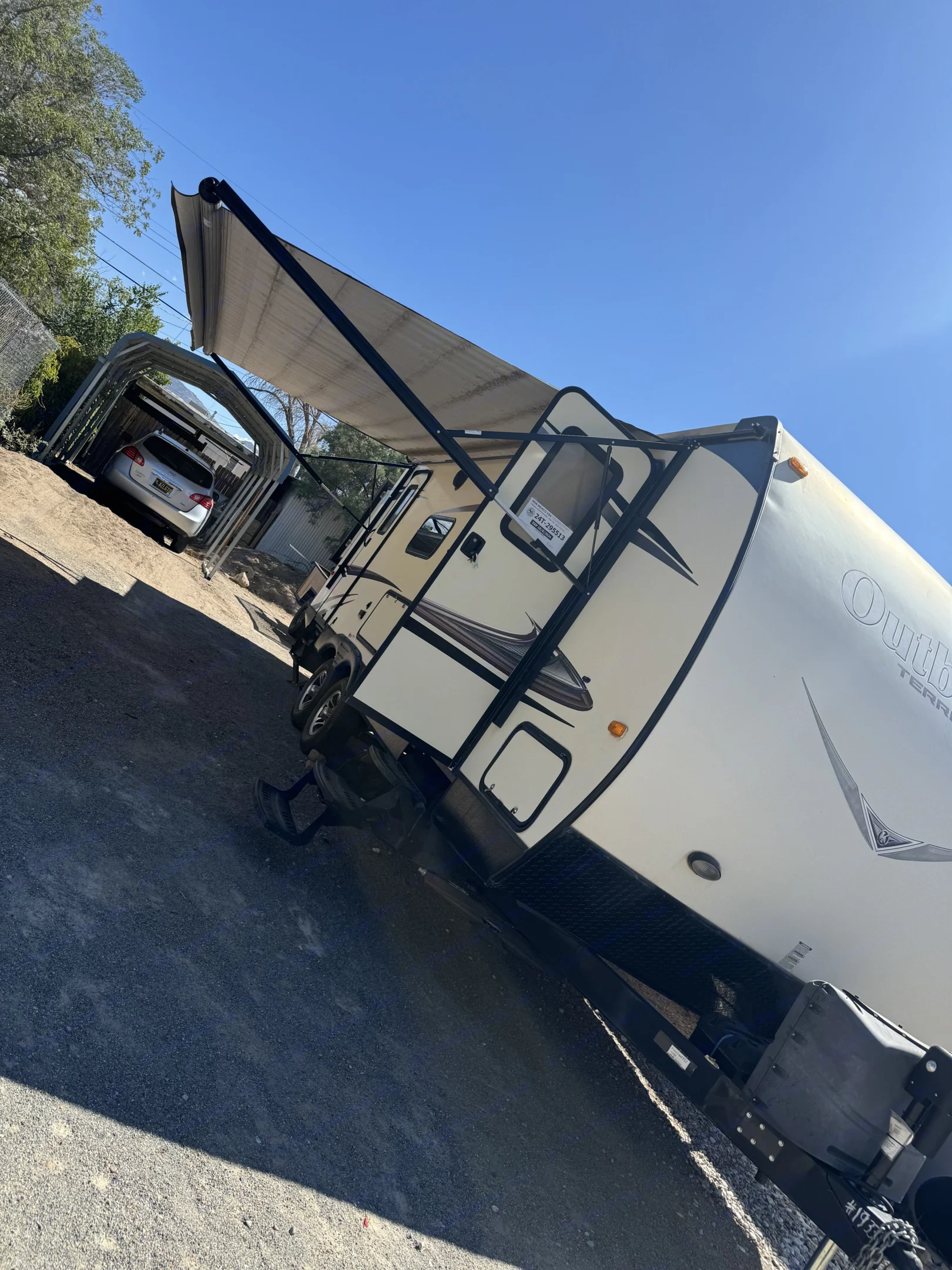 An RV rental, cream-colored with its awning extended, parked on gravel next to a car under a carport.. Keystone RV Outback Terrain 2014