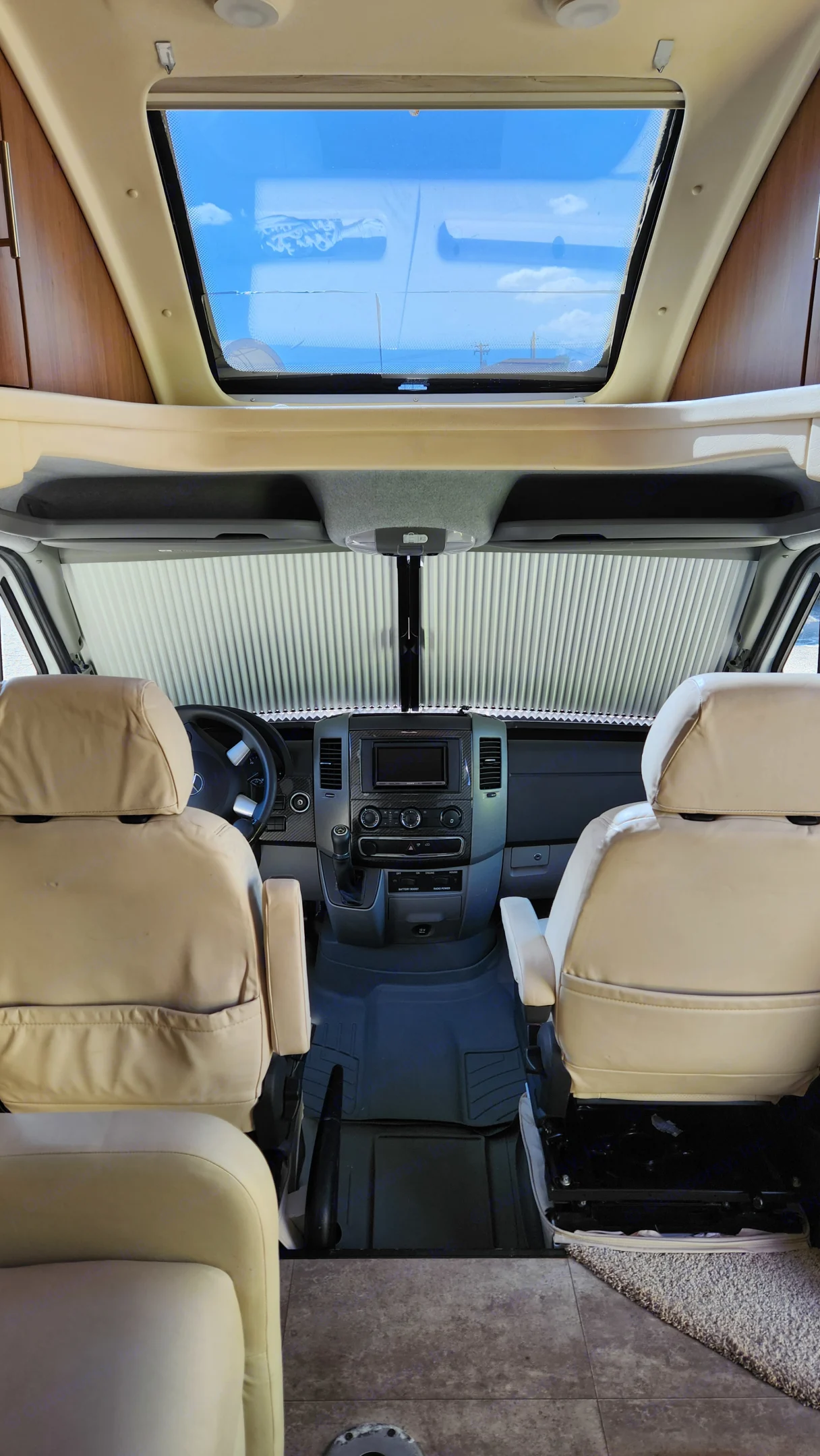 RV rental interior. View from the living area into the bright cockpit, featuring tan seats, dashboard, sunshade, and an overhead skylight showing blue sky.. Winnebago View 2015