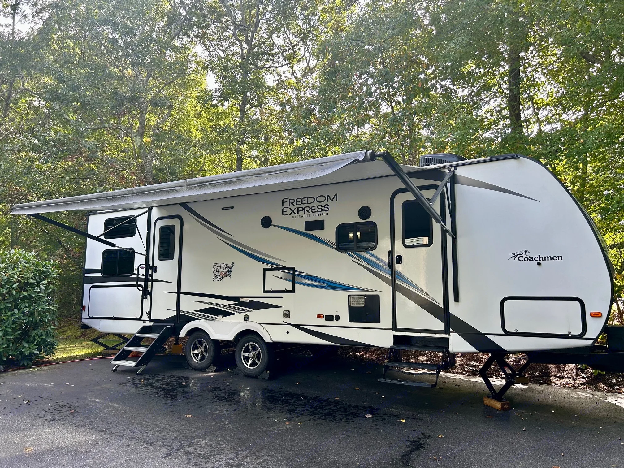 A white Coachmen Freedom Express travel trailer RV rental, with its awning extended, parked on asphalt with green trees in the background.. Coachmen Freedom Express 287 BHDS 2021