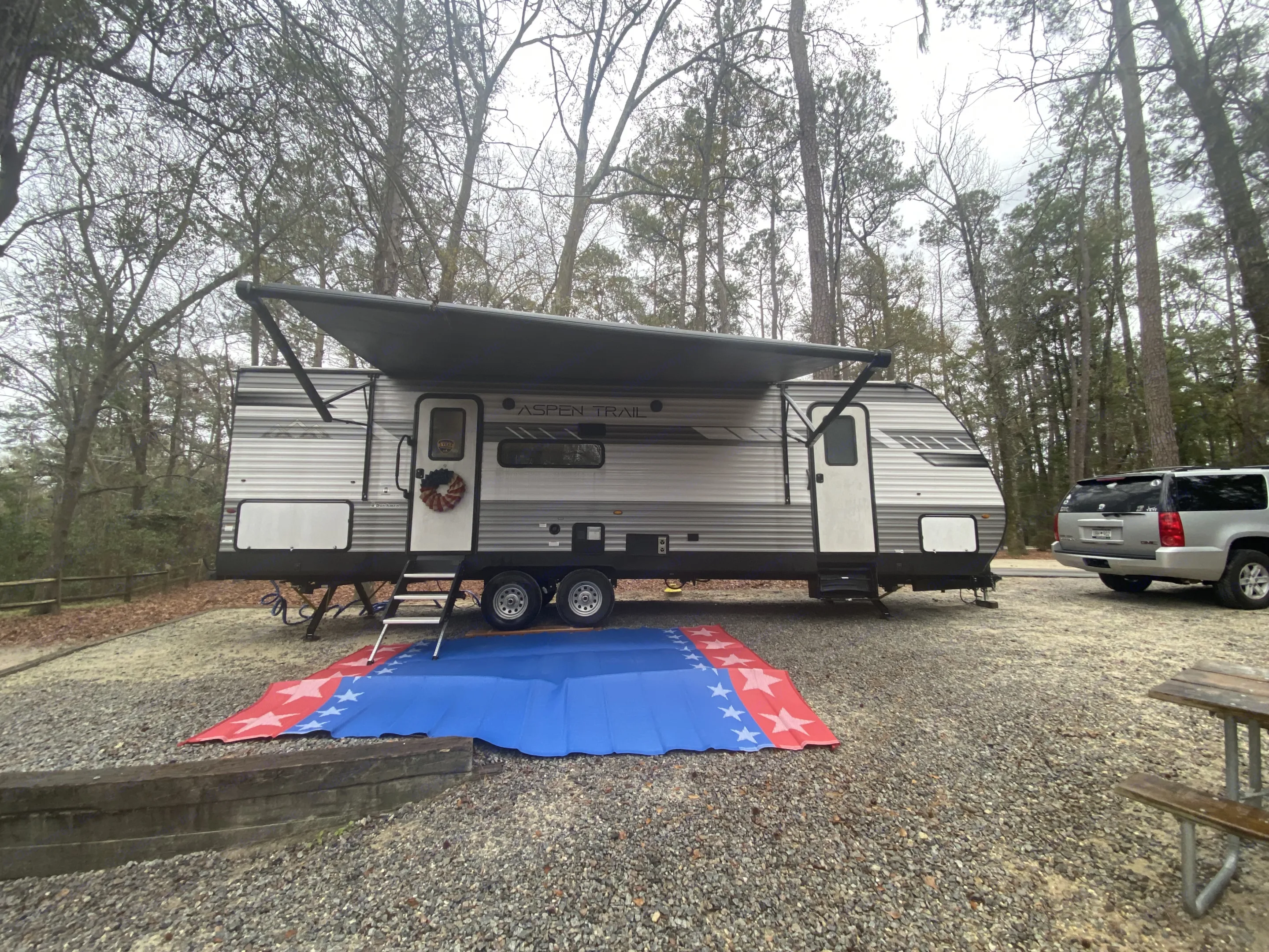 An RV rental with an extended awning is set up on a wooded gravel campsite, featuring a red and blue star mat.. Dutchmen Aspen Trail 2021