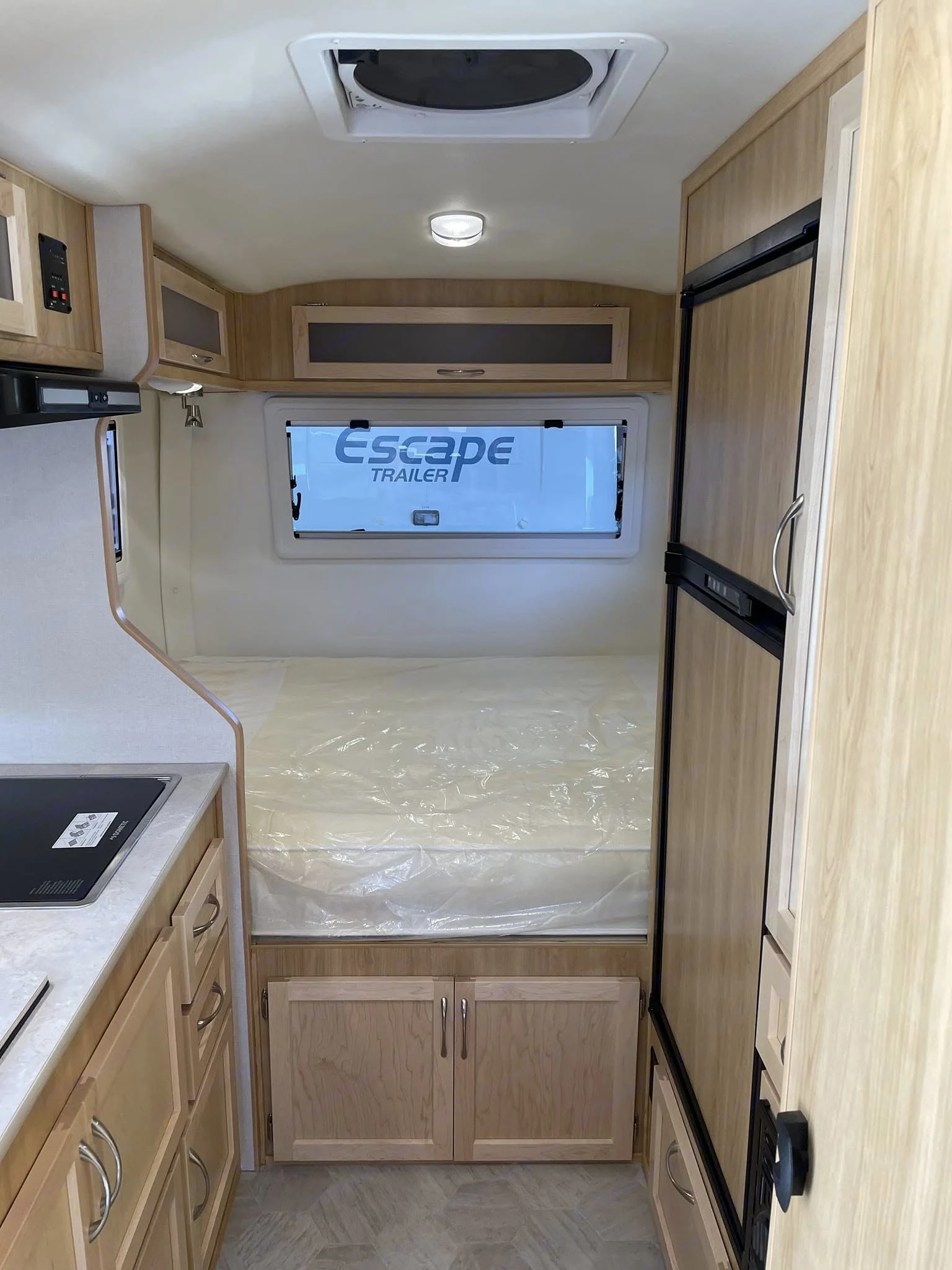 Compact rv rental interior: kitchen on left, bed with plastic cover at back, fridge on right. Light-wood finish.. Escape 19 2024