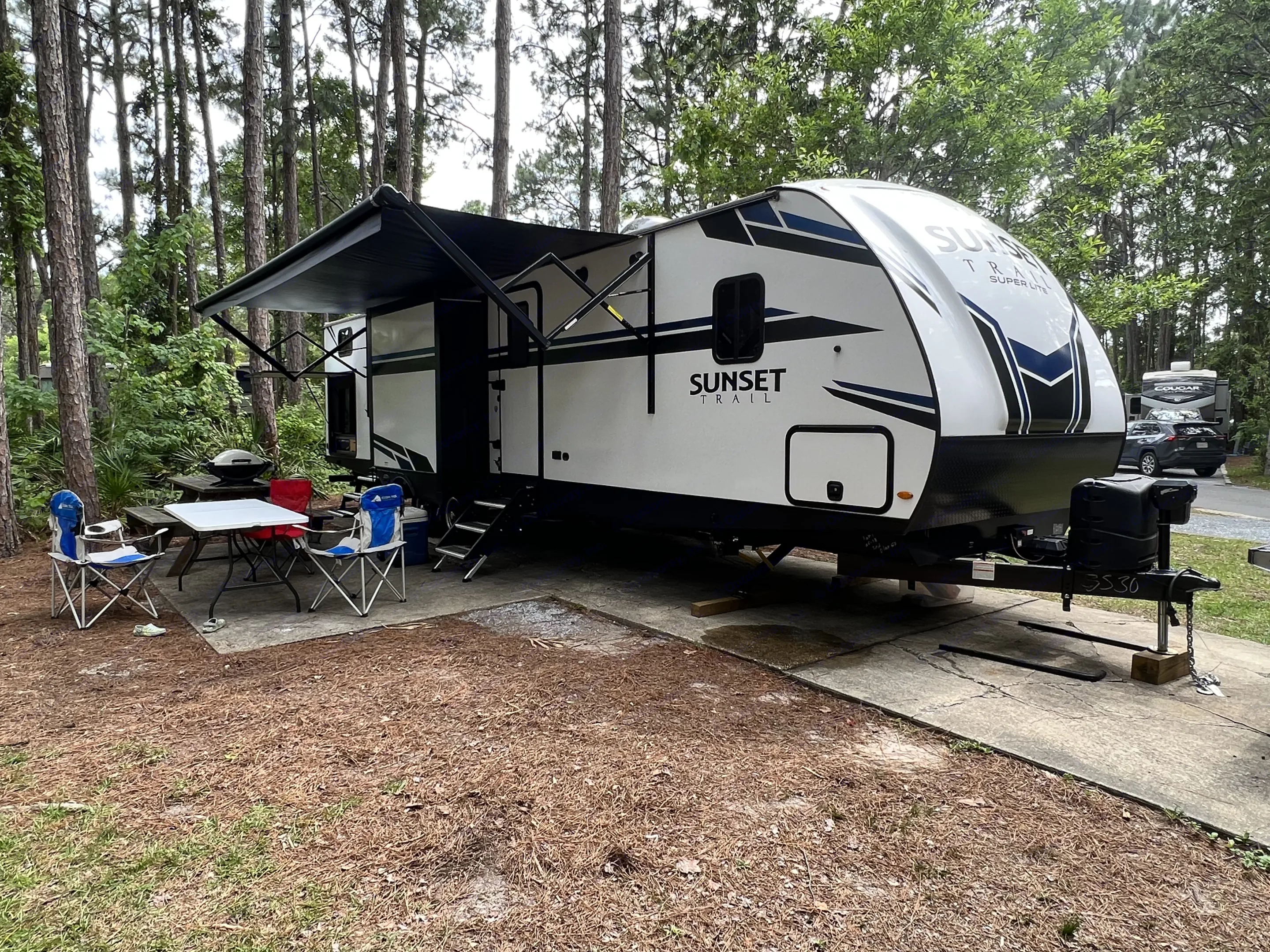 A white travel trailer RV rental, with an extended awning, parked at a wooded campsite with a picnic area.. Crossroads RV Sunset Trail Super Lite 2021