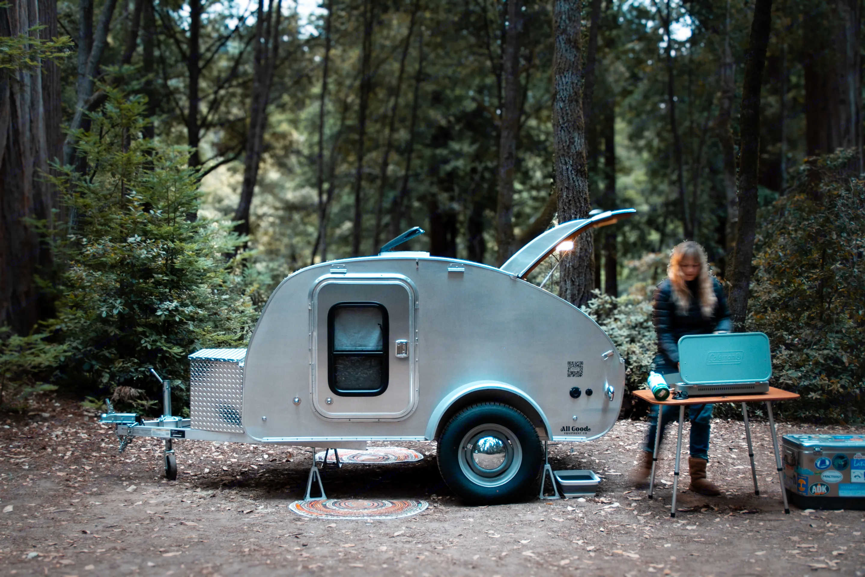 Teardrop RV rental at a forest campsite, with a person setting up a Coleman stove for cooking. Ideal for outdoor adventures.. All Good Equipment Company Teardrop Trailer 2024