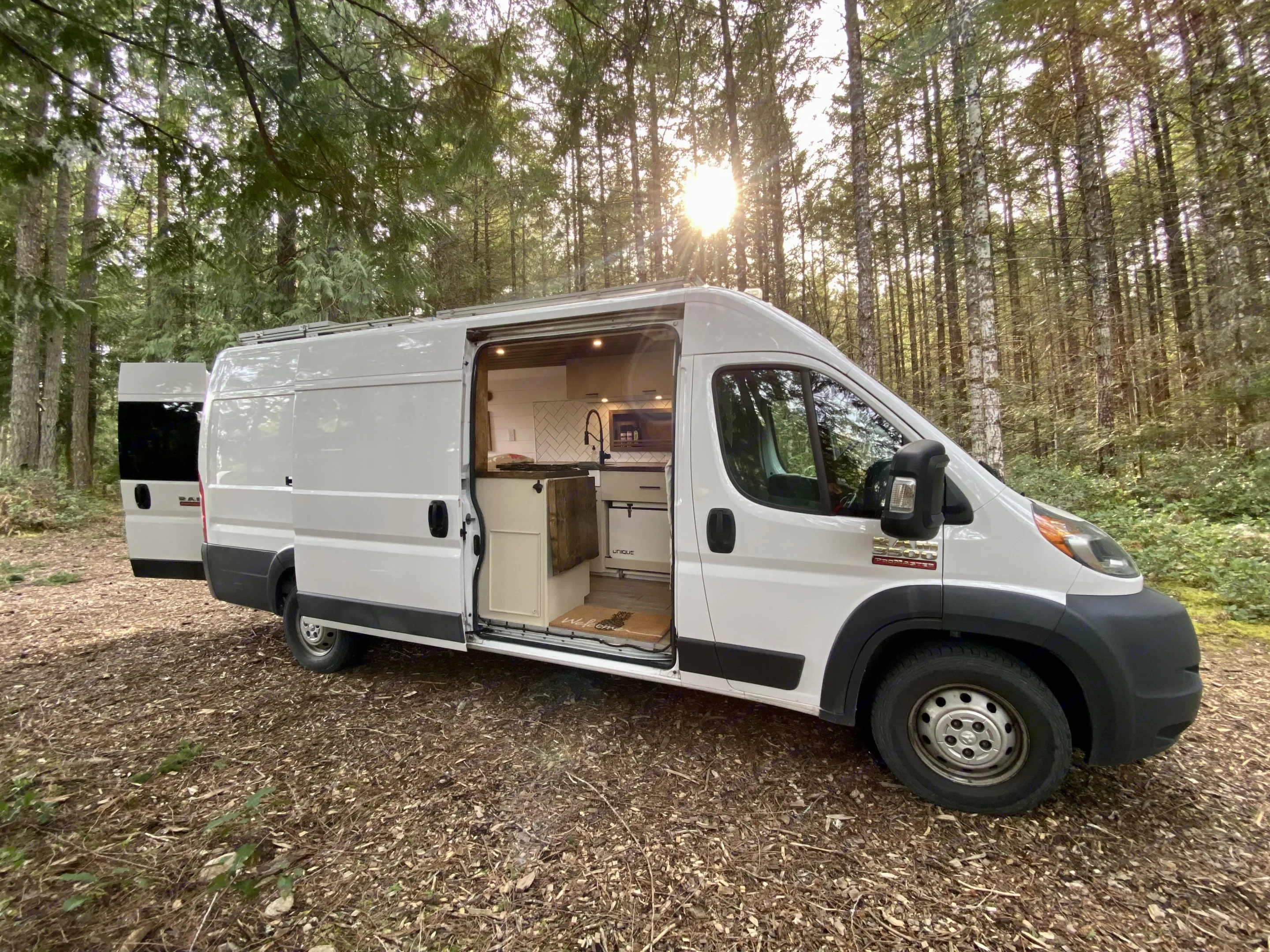 White RV rental camper van in sunny forest, doors open to reveal detailed kitchen & cozy interior. Great for van life adventures!. Ram Promaster 2018