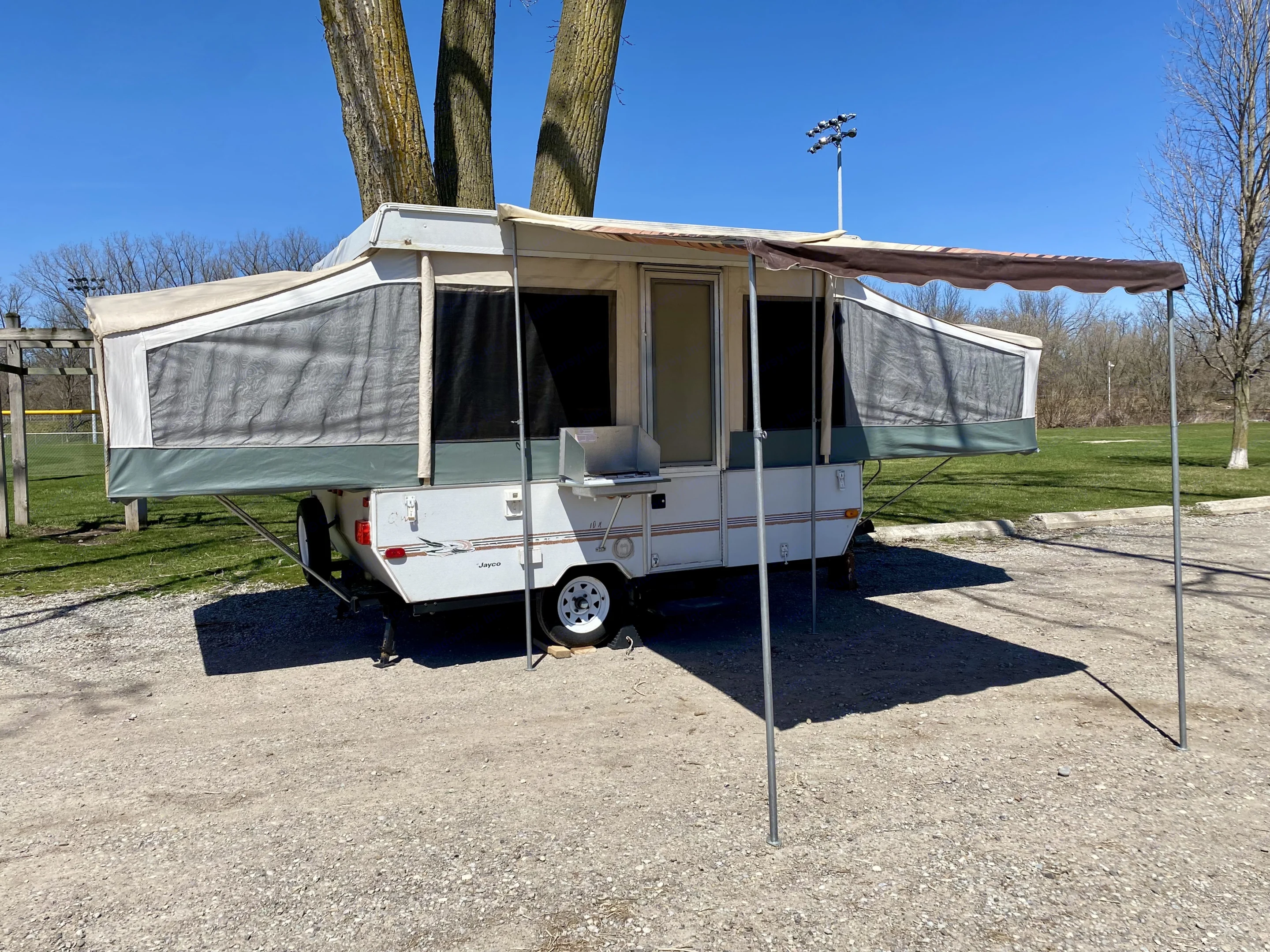 Pop-up RV rental set up at a sunny campground with an awning and outdoor stove, ready for adventure.. Jayco Qwest 2000