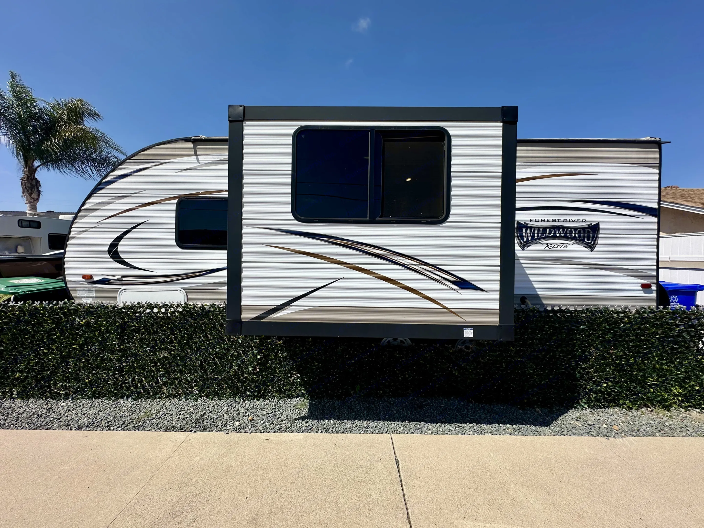 A white RV with a slide-out extended, parked behind a lush green hedge and gravel. Ready for your next rv rental trip under a clear blue sky.. Forest River Wildwood X-Lite 2015