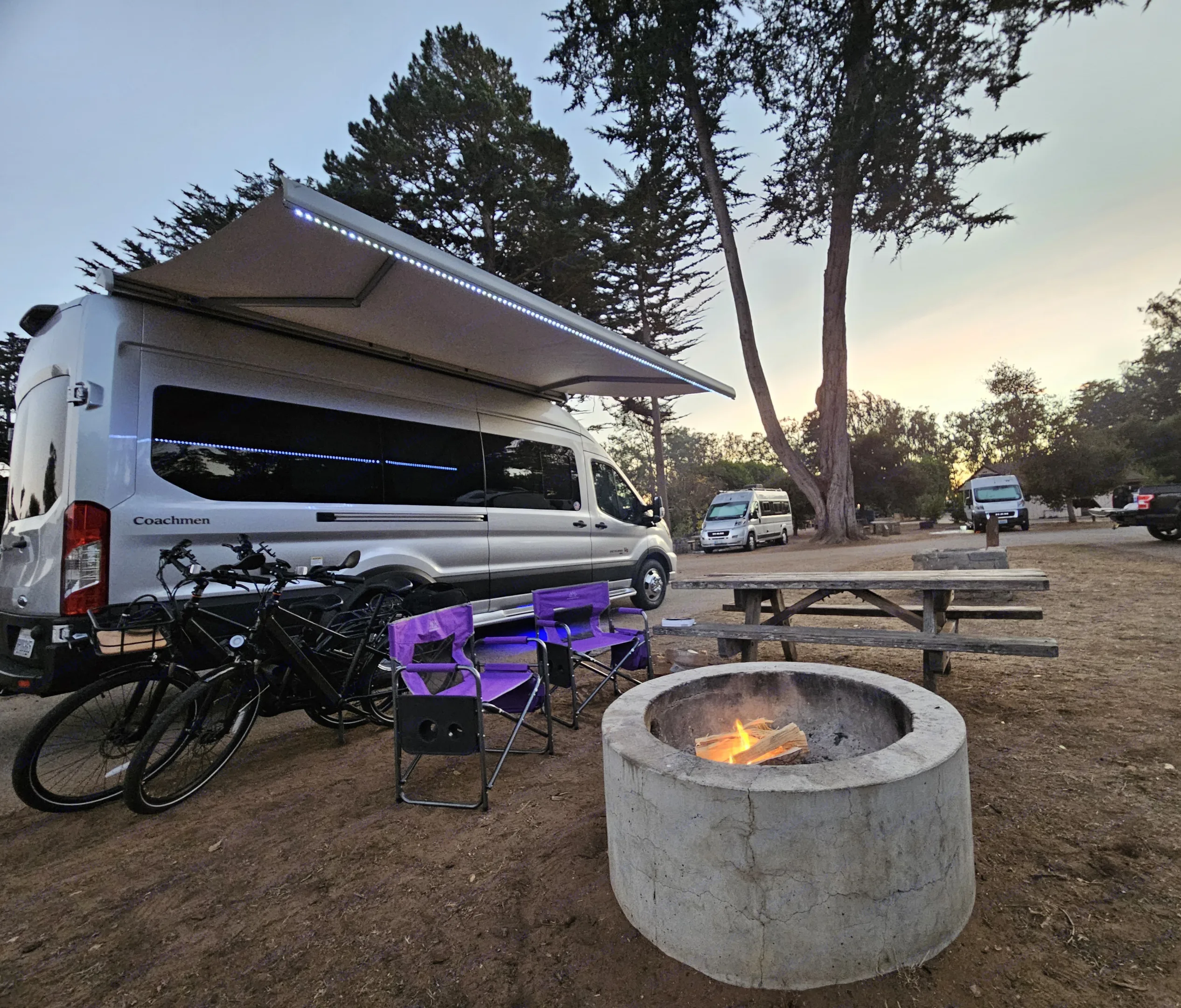 Relaxing campsite evening: a bright RV rental campervan with awning, bikes, chairs, and a fire pit glowing at dusk.. Forest River Coachmen Beyond 2023