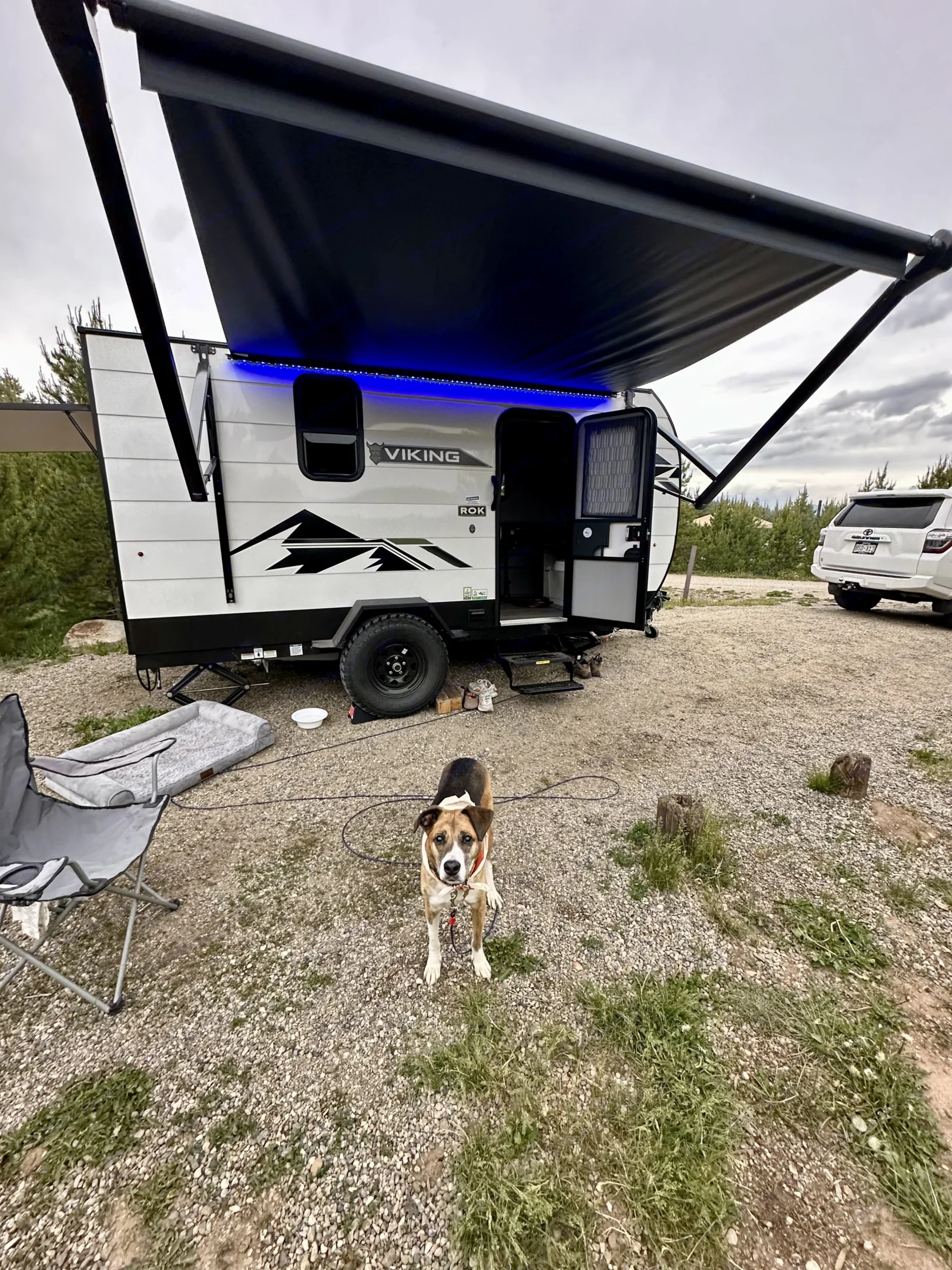 Bright white Viking RV rental, with cool blue LED awning lights, parked on a gravel campsite. A friendly dog stands in front, ready for fun.. Forest River Viking 12000 2024