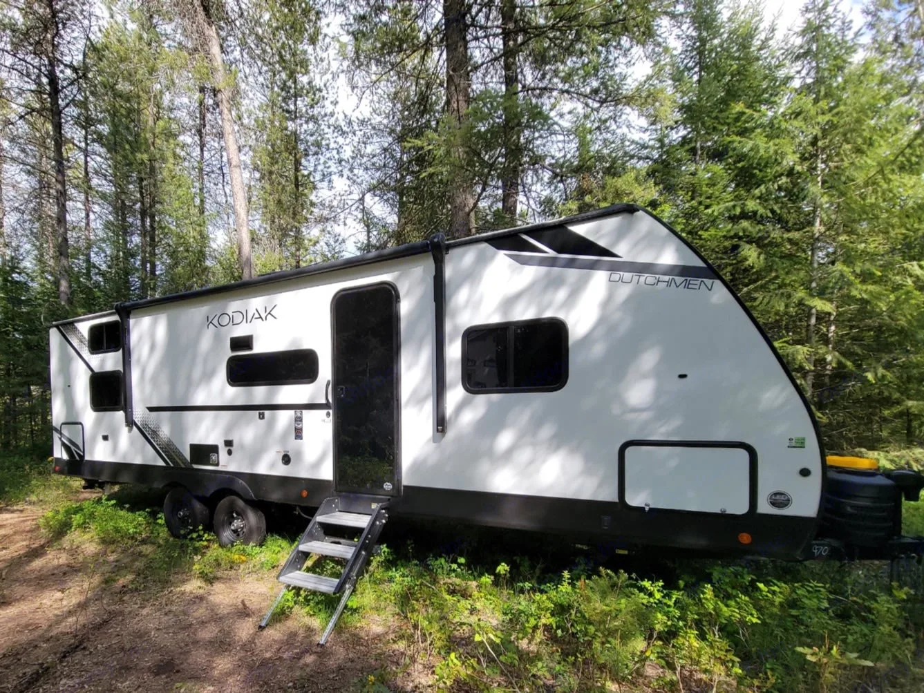 White Kodiak Dutchmen travel trailer RV rental parked in a green forest campsite with steps deployed. Ideal for camping outdoors.. Dutchmen Kodiak 2025
