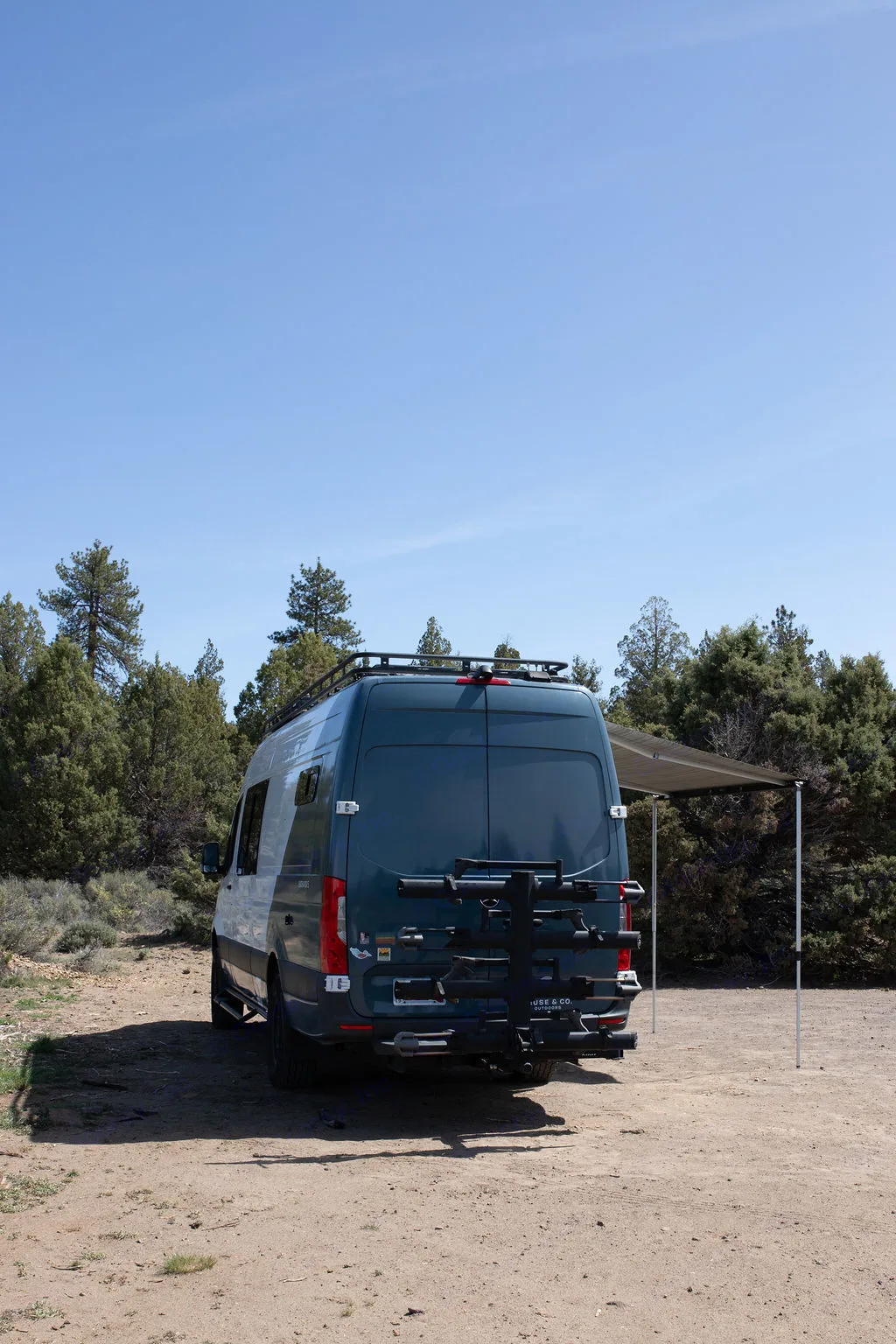 Dark blue Sprinter van RV rental, rear view, with roof rack & awning extended in a dirt clearing among. Mercedes-Benz Sprinter 2020