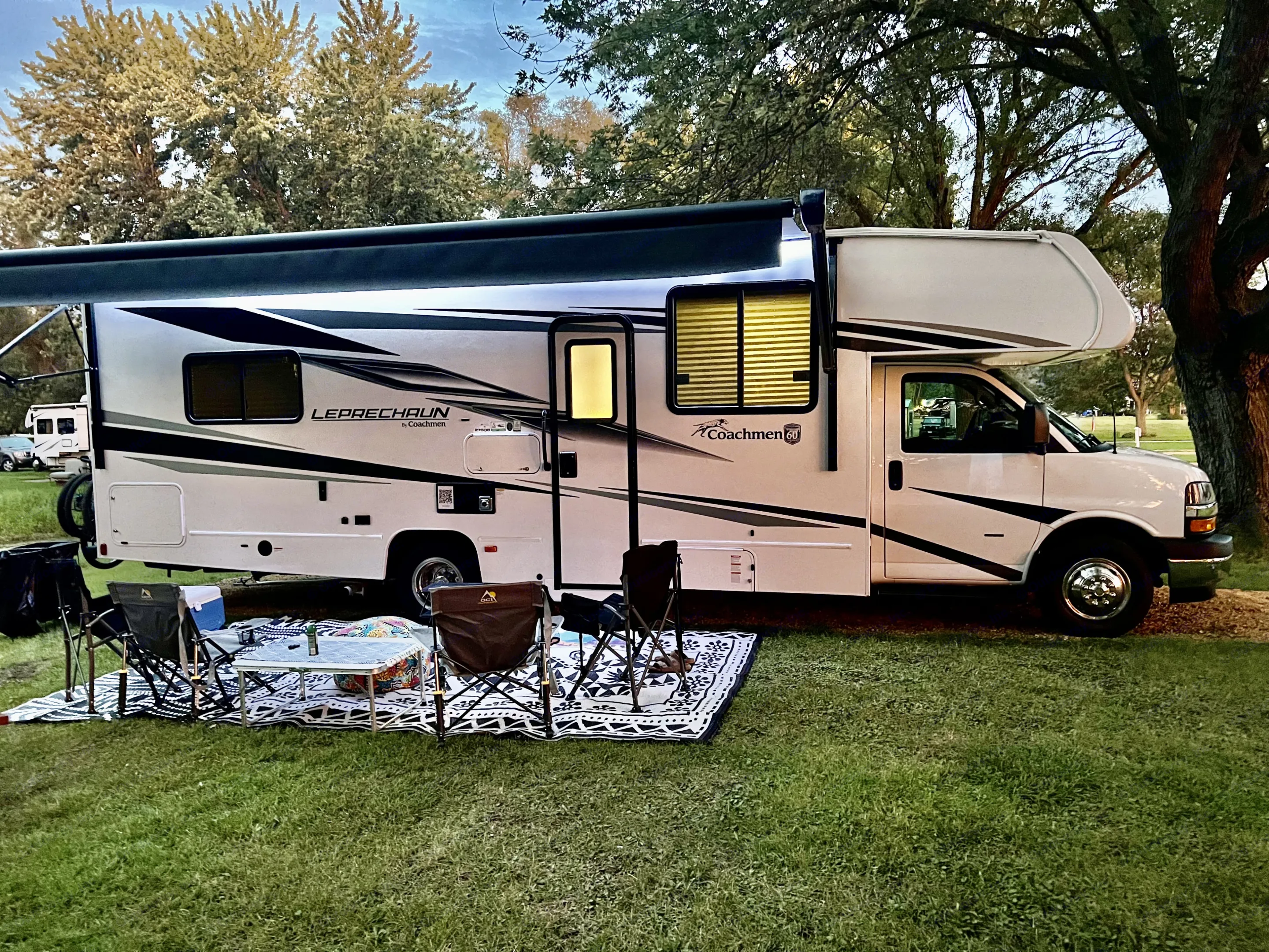 White Coachmen Leprechaun RV rental set up at a grassy campsite with awning, outdoor chairs, table, and rug for relaxing.. Forest River Coachmen Leprechaun 2024