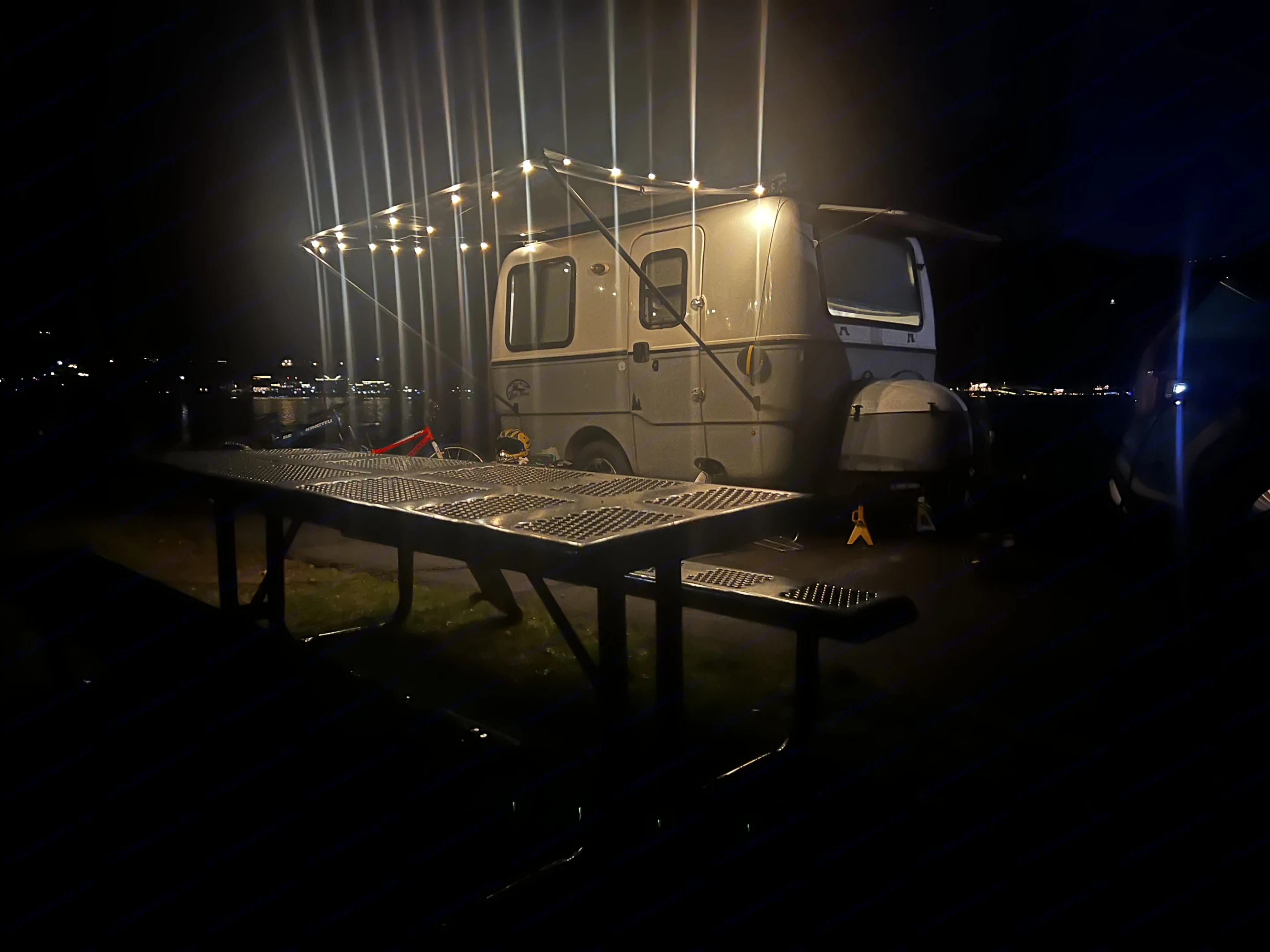 Illuminated RV rental setup at night with a picnic table, string lights, and distant town lights across water.. Outback Trillium 2020