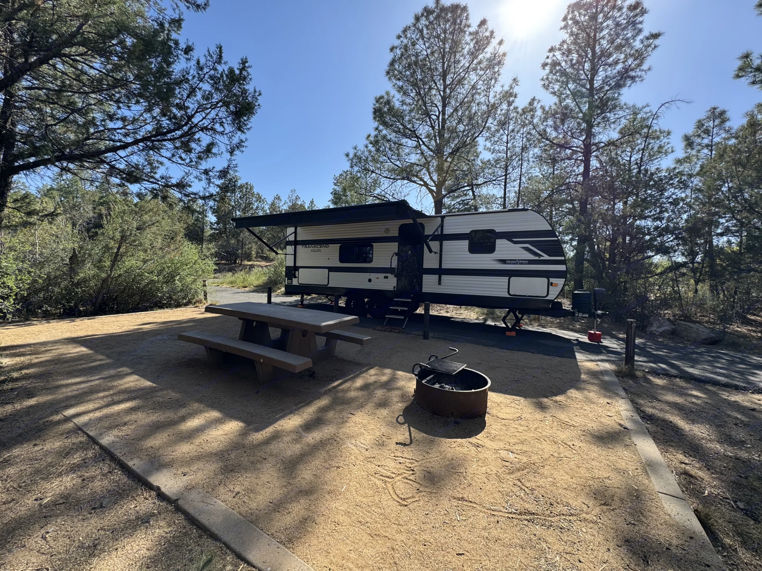 An RV rental travel trailer is set up at a sunny campsite with a picnic table and fire pit, surrounded by tall trees.. Grand Design Transcend Xplor 26BHX 2024