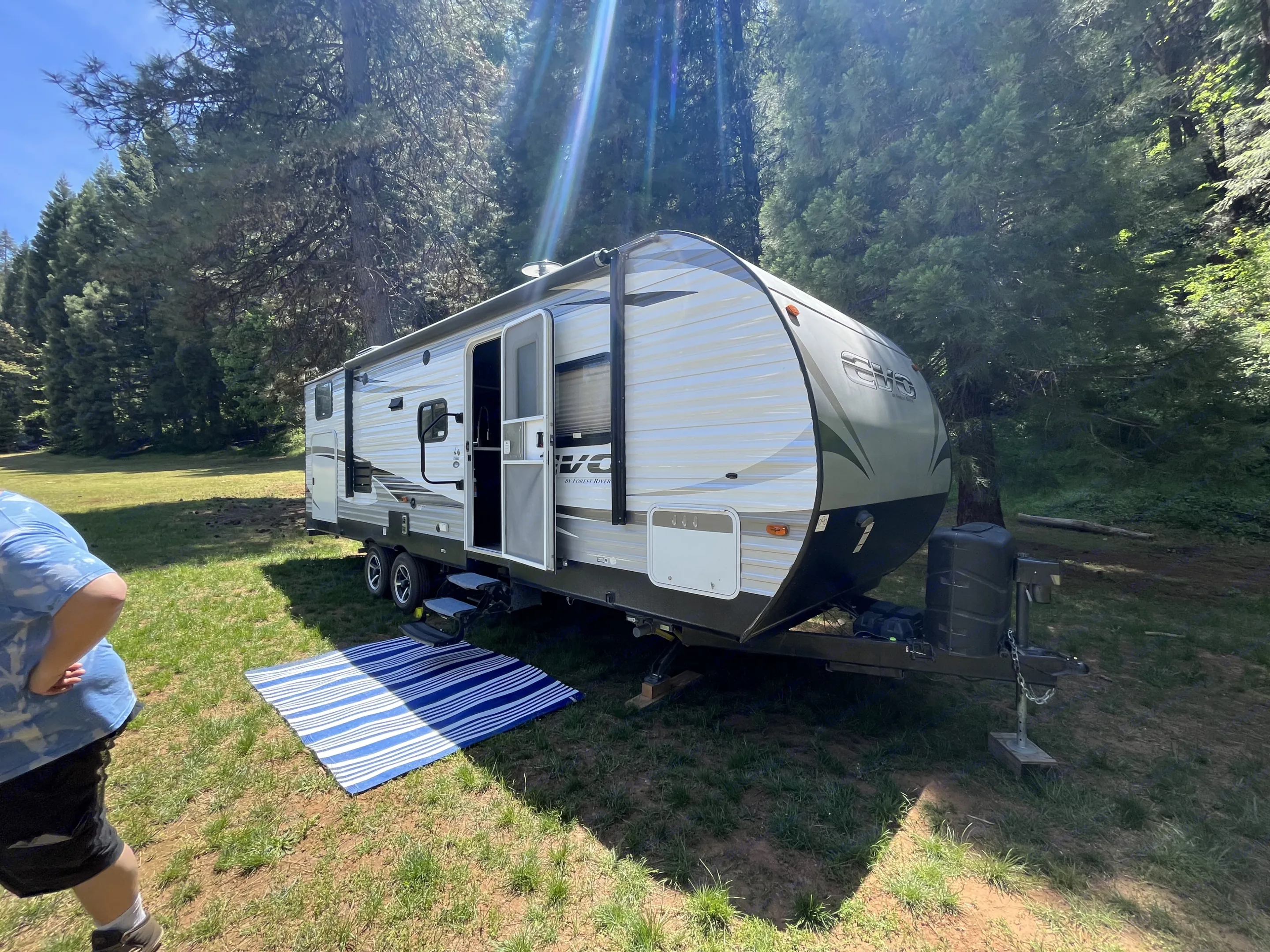 An RV rental trailer is parked on a grassy site amidst tall forest trees, with a blue mat inviting outdoor relaxation.. Forest River Flagstaff 2018