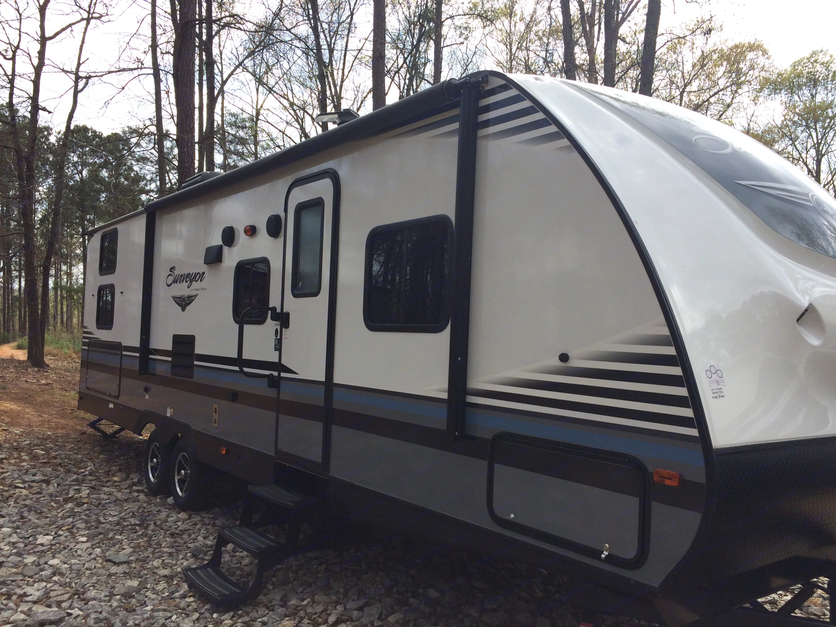 This white & gray Surveyor travel trailer, parked on gravel with trees, is ready for your next rv rental adventure.. Forest River Surveyor 2017