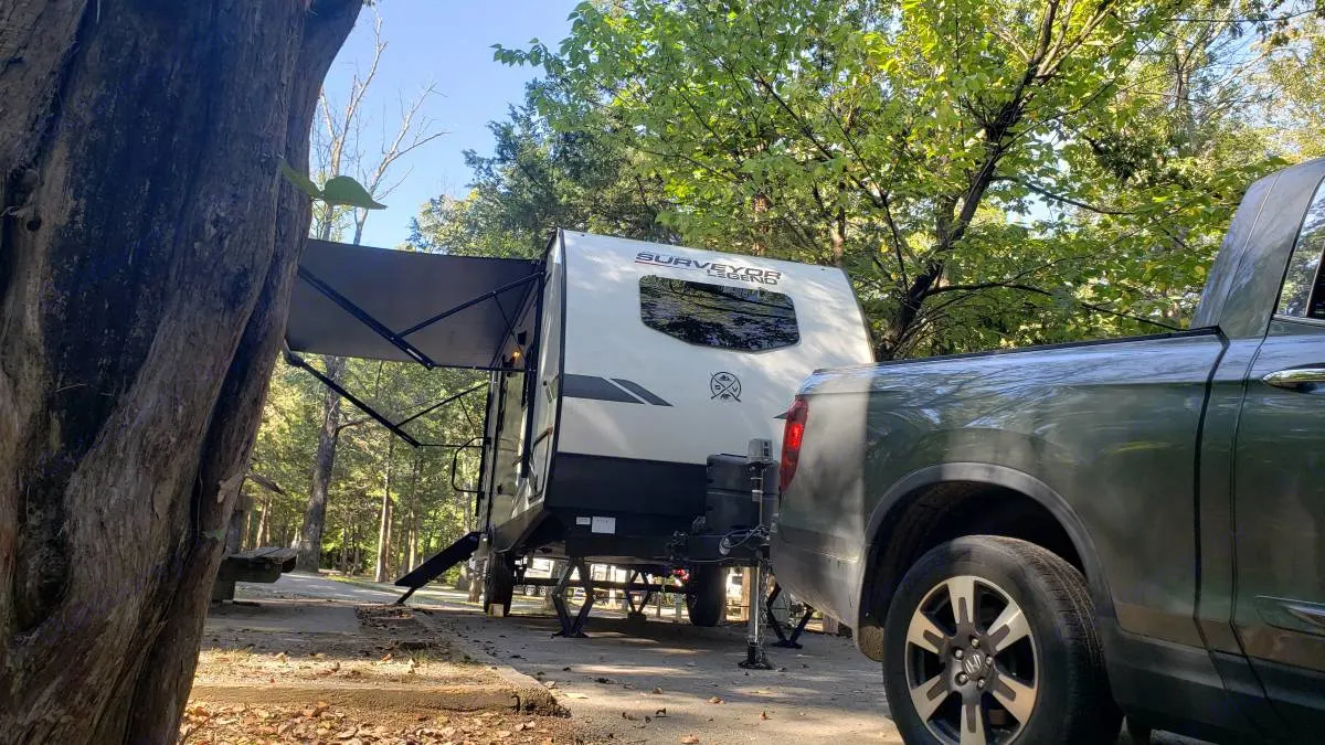 A white Surveyor Legend RV rental, with awning out, is hitched to a pickup truck at a leafy, shaded campsite.. Forest River Surveyor Legend 19RBLE 2022