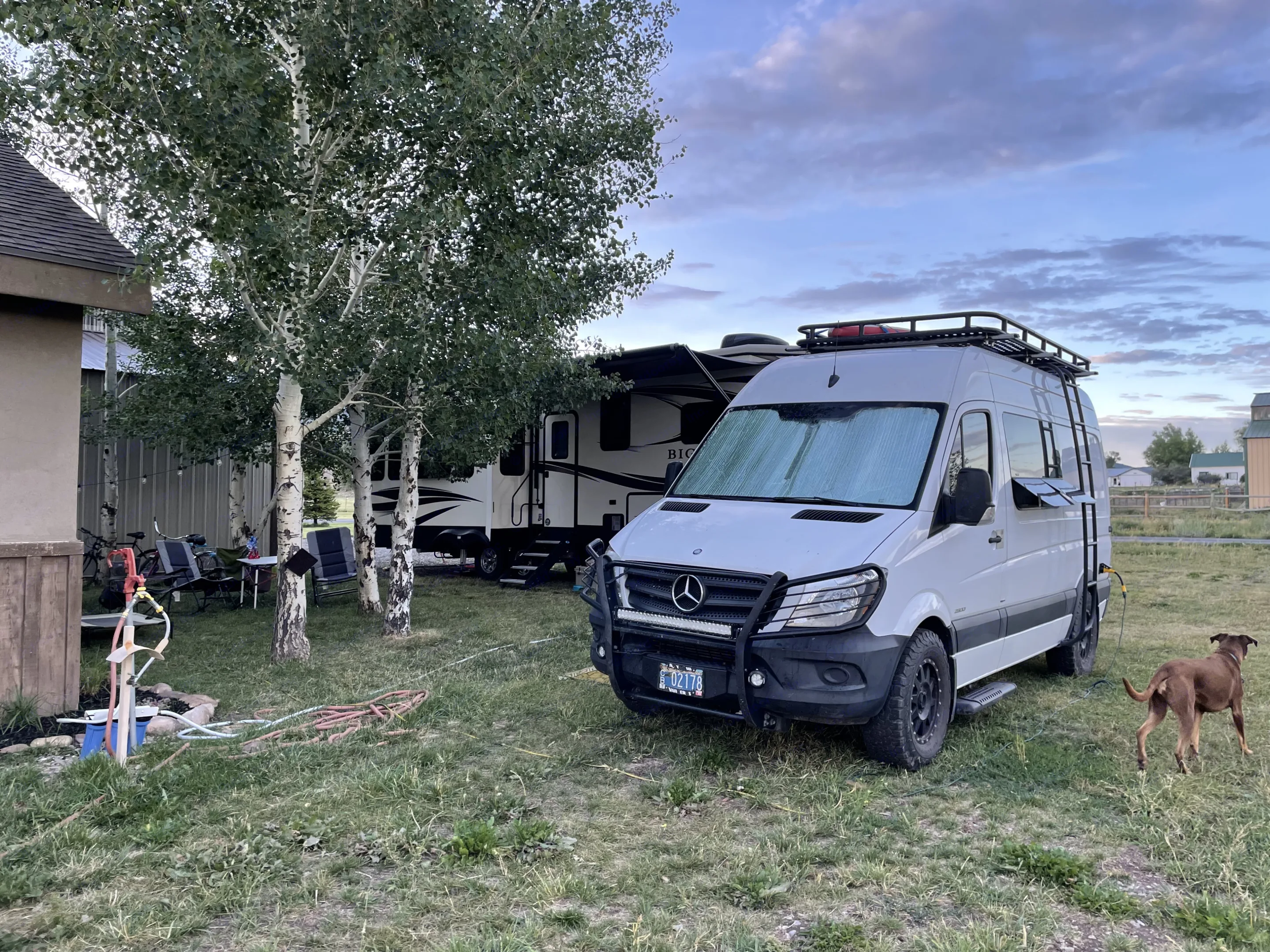 A white Mercedes Sprinter van RV rental and a large travel trailer set up in a grassy campsite with aspen trees and a dog.. Mercedes-Benz Sprinter 2015