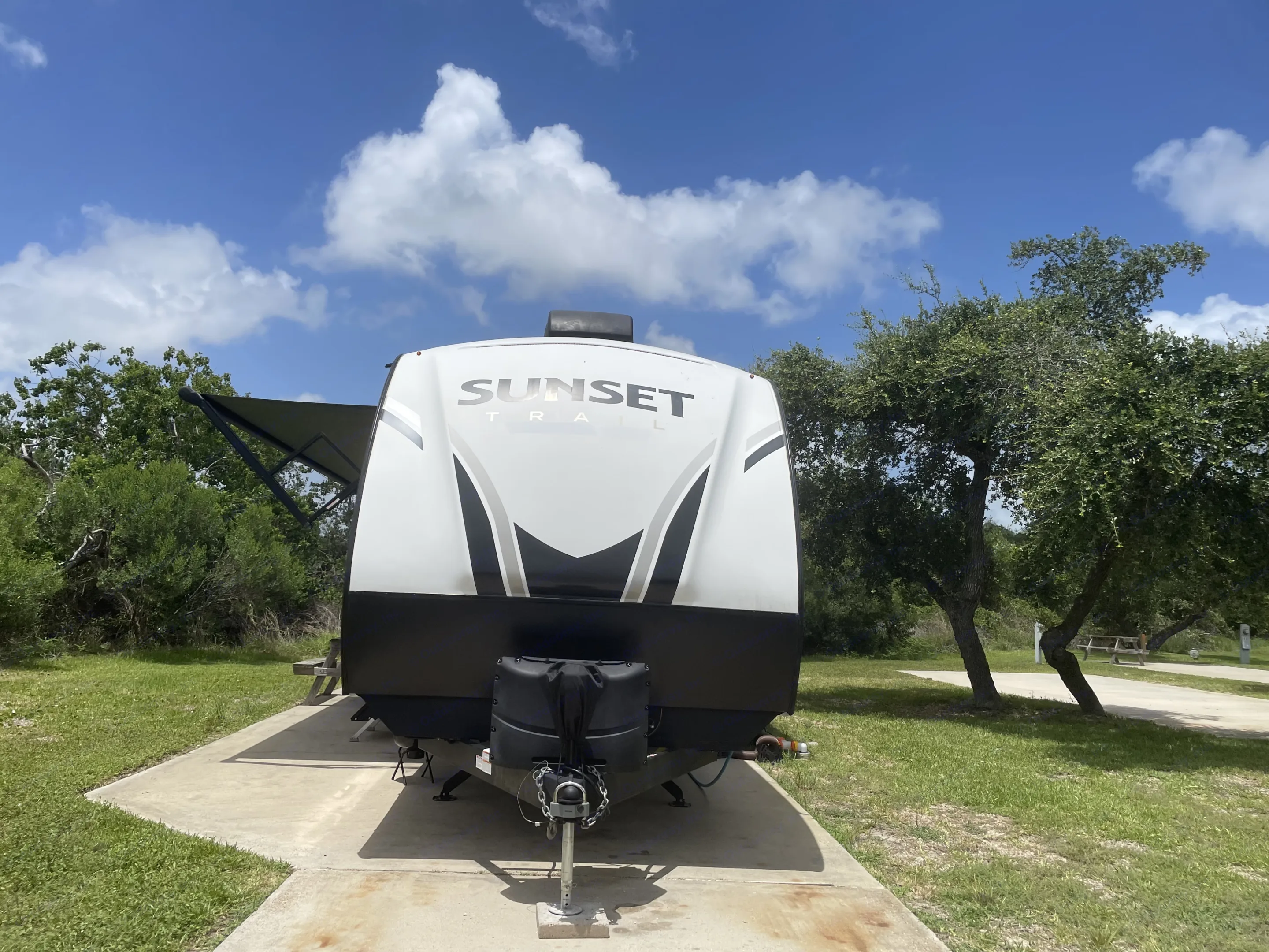 A 'Sunset Trail' RV rental is parked on a concrete pad at a sunny campsite, surrounded by green grass and trees.. Crossroads RV Sunset Trail Super Lite 2023