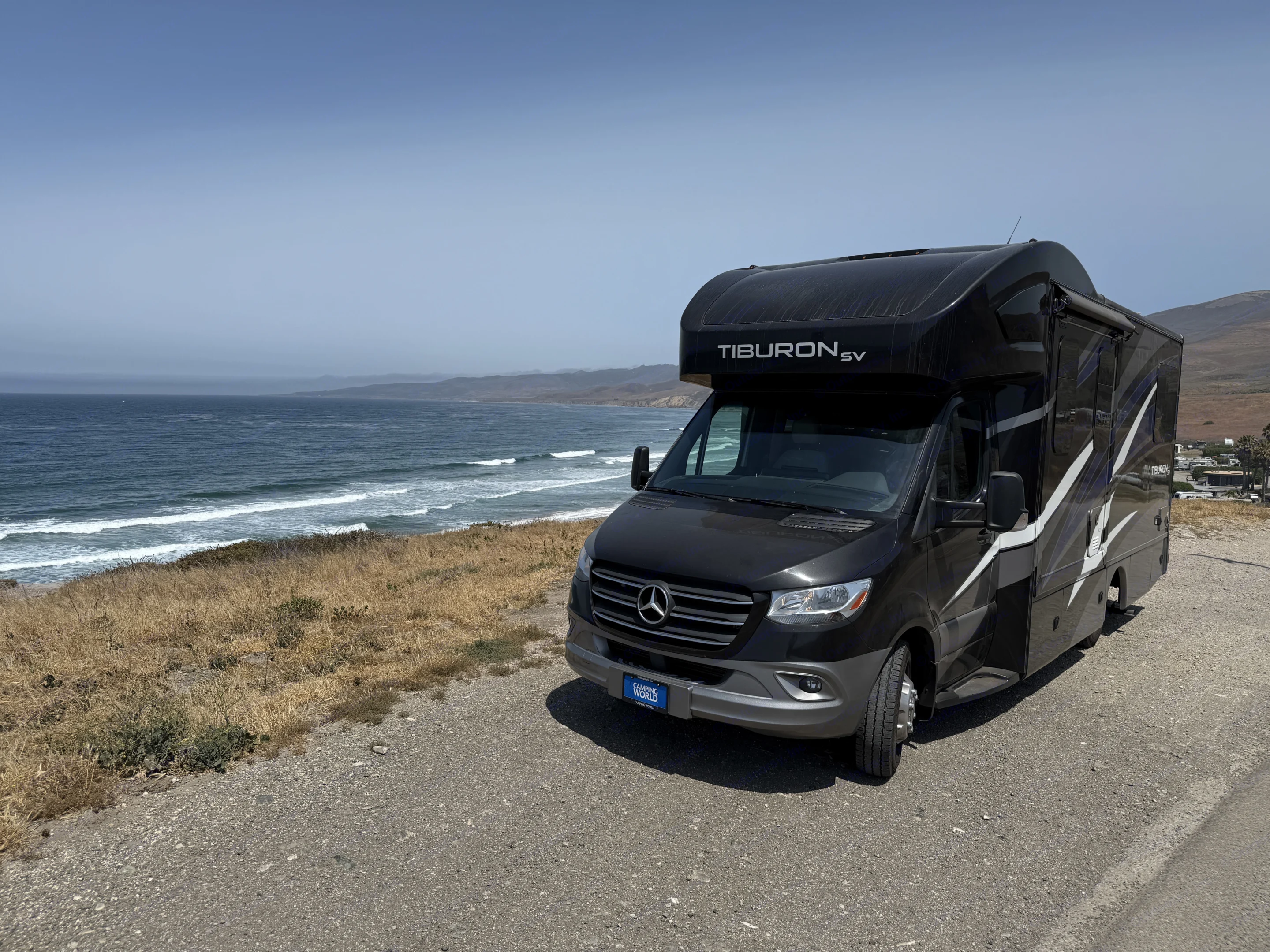 A black Tiburon SV RV rental is parked on a gravel road overlooking the blue ocean and coastal hills.. Thor Tiburon 2022