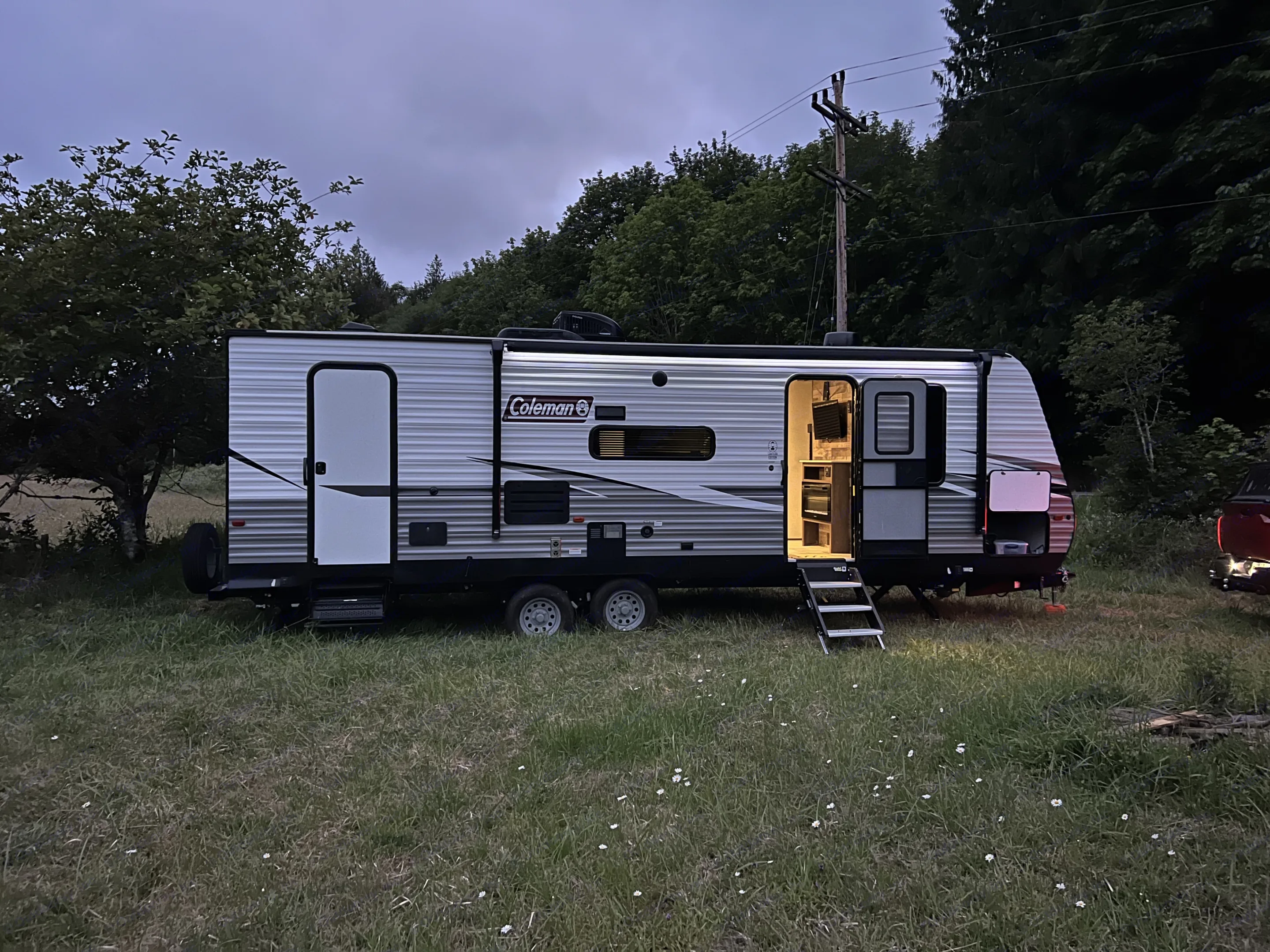 Evening RV rental setup in a grassy field. The Coleman travel trailer is lit up with steps down, ready for camping.. Dutchmen Coleman Lantern 2021