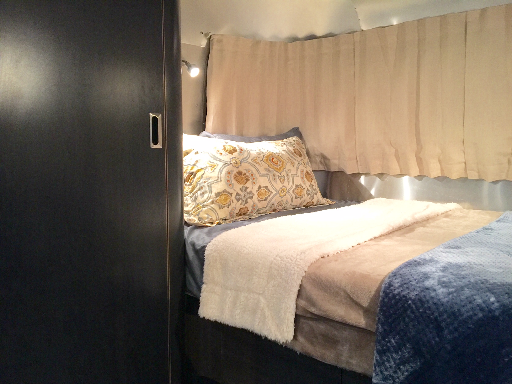 Cozy RV rental bedroom featuring a patterned pillow, layered blankets, a reading light, and distinct metallic walls with beige curtains.. Airstream International 2003