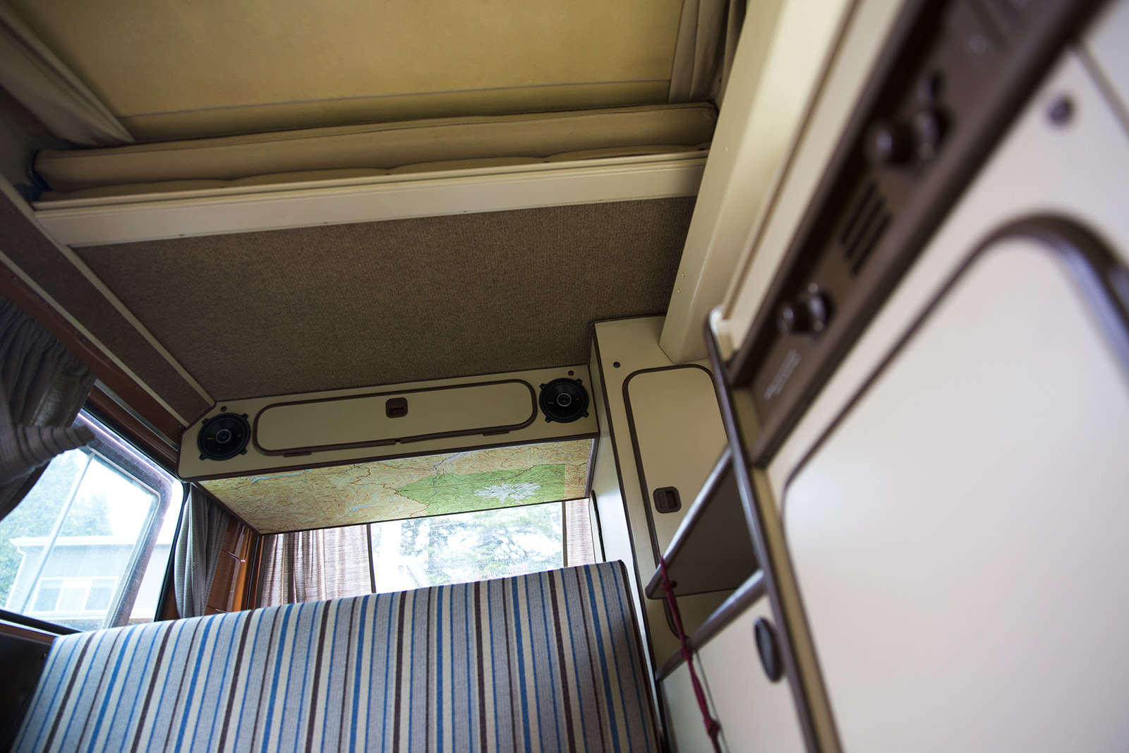 Cozy RV rental camper van interior featuring a raised pop-top bed, map headliner, striped bench, and storage.. Volkswagen Vanagon 1983