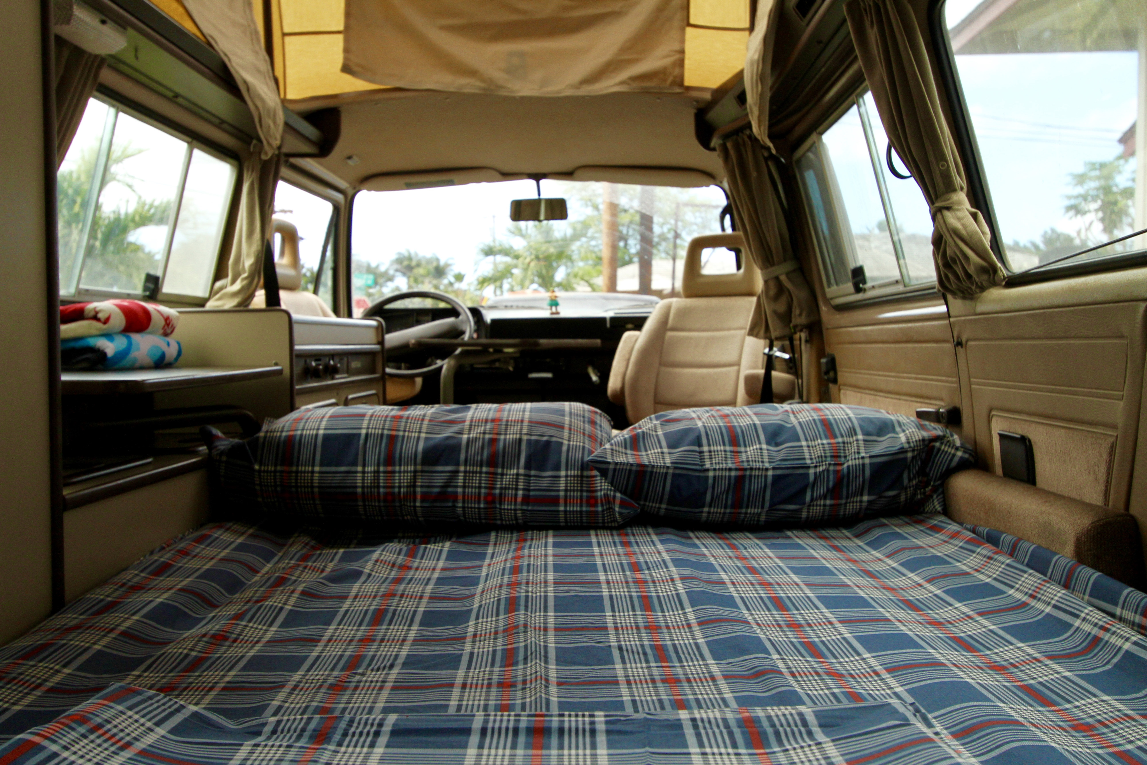 Cozy RV rental interior: blue plaid bed, towels on shelf, and front view of steering wheel and tropical outdoors.. Volkswagen Vanagon 1984