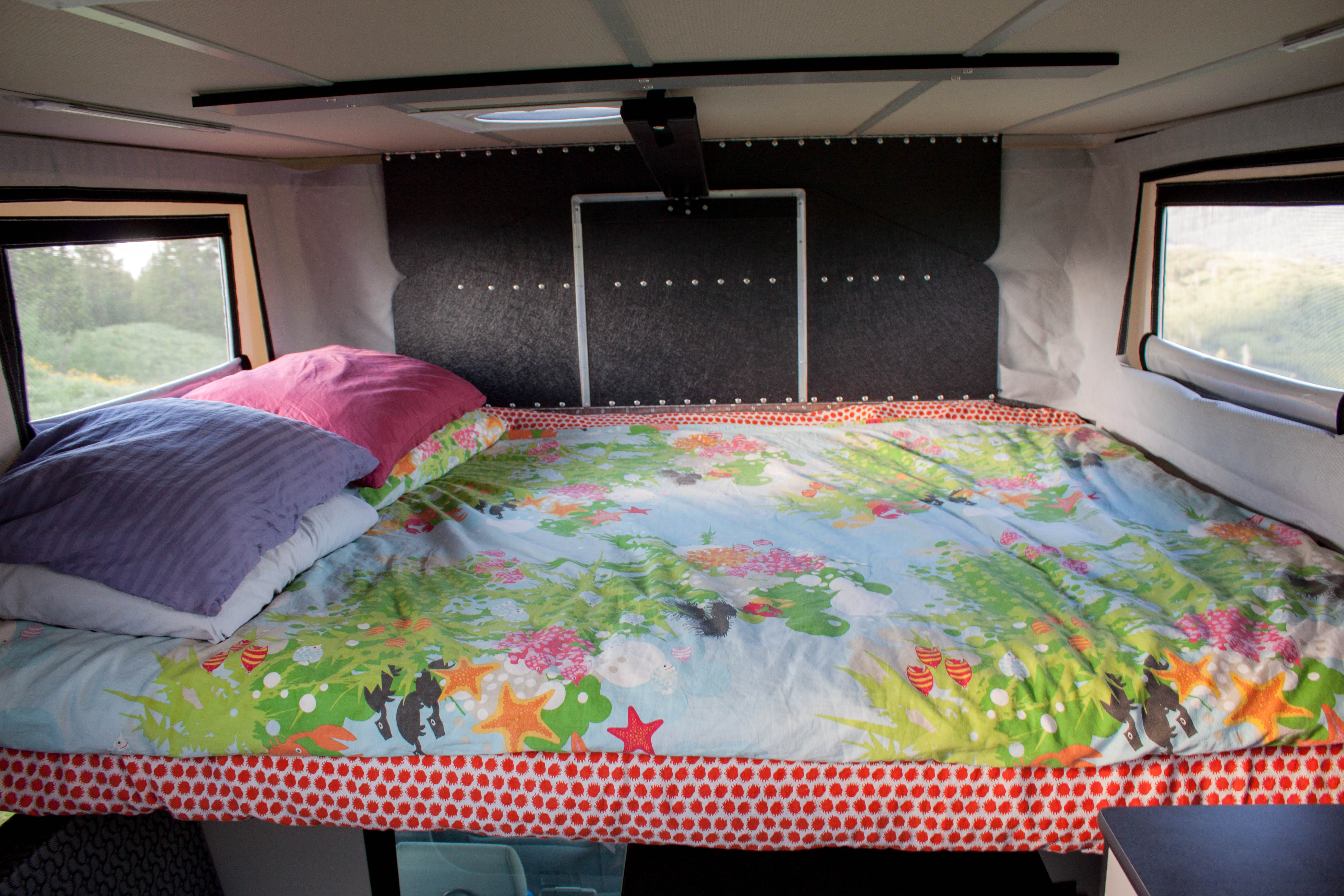 Inside an RV rental: a comfy bed with vibrant bedding, pillows, and windows offering scenic nature views.. Chevrolet Silverado 2016