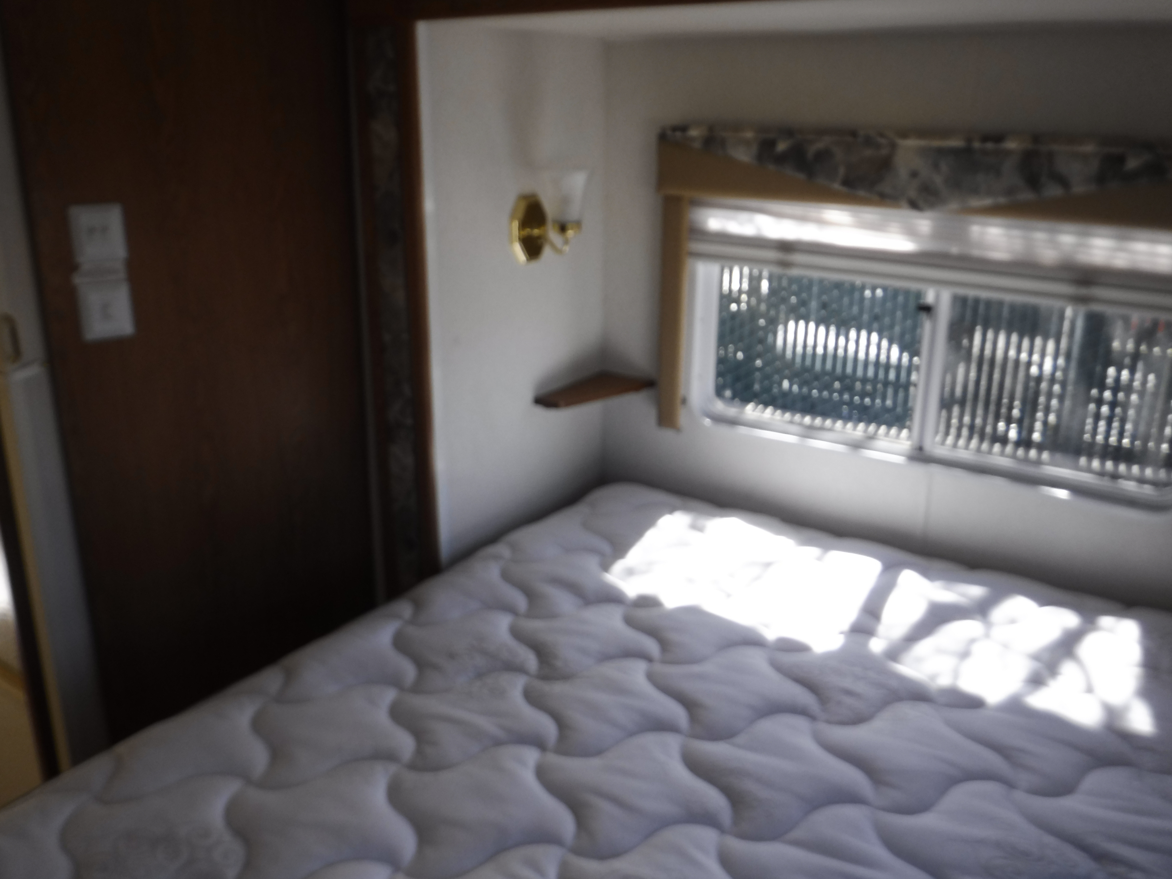 RV rental bedroom interior: shows a quilted mattress, bright window, wall light, and small wooden shelf.. HOLIDAY RAMBLER ATLANTIS 2002