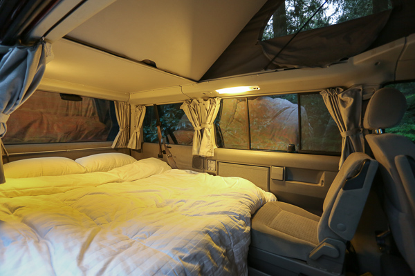 Cozy RV rental interior featuring a large bed with white linens, illuminated by warm lights. Outside, forest views through the windows.. Volkswagen Eurovan weekender 2003