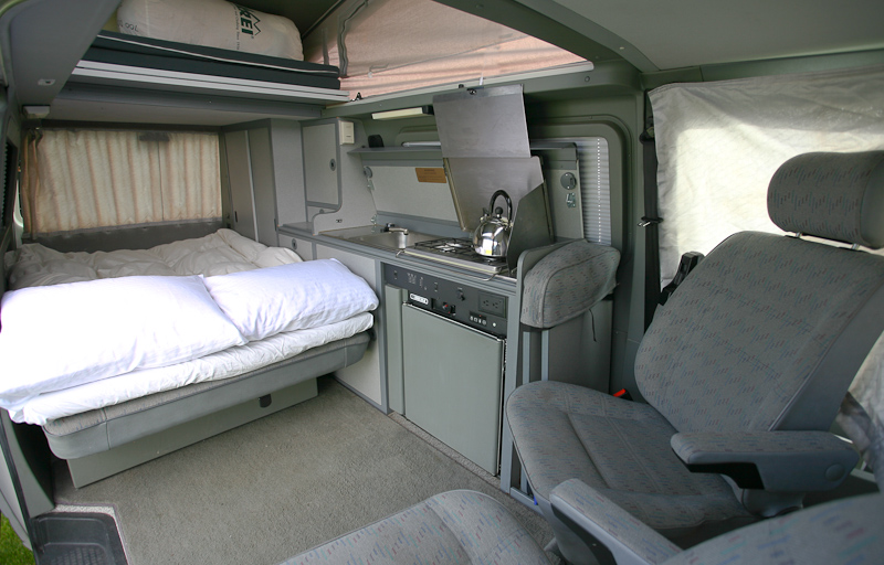 Spacious RV rental interior featuring a cozy bed, compact kitchen with kettle, and front seats. Ideal for travel.. Volkswagen Eurovan camper 2000