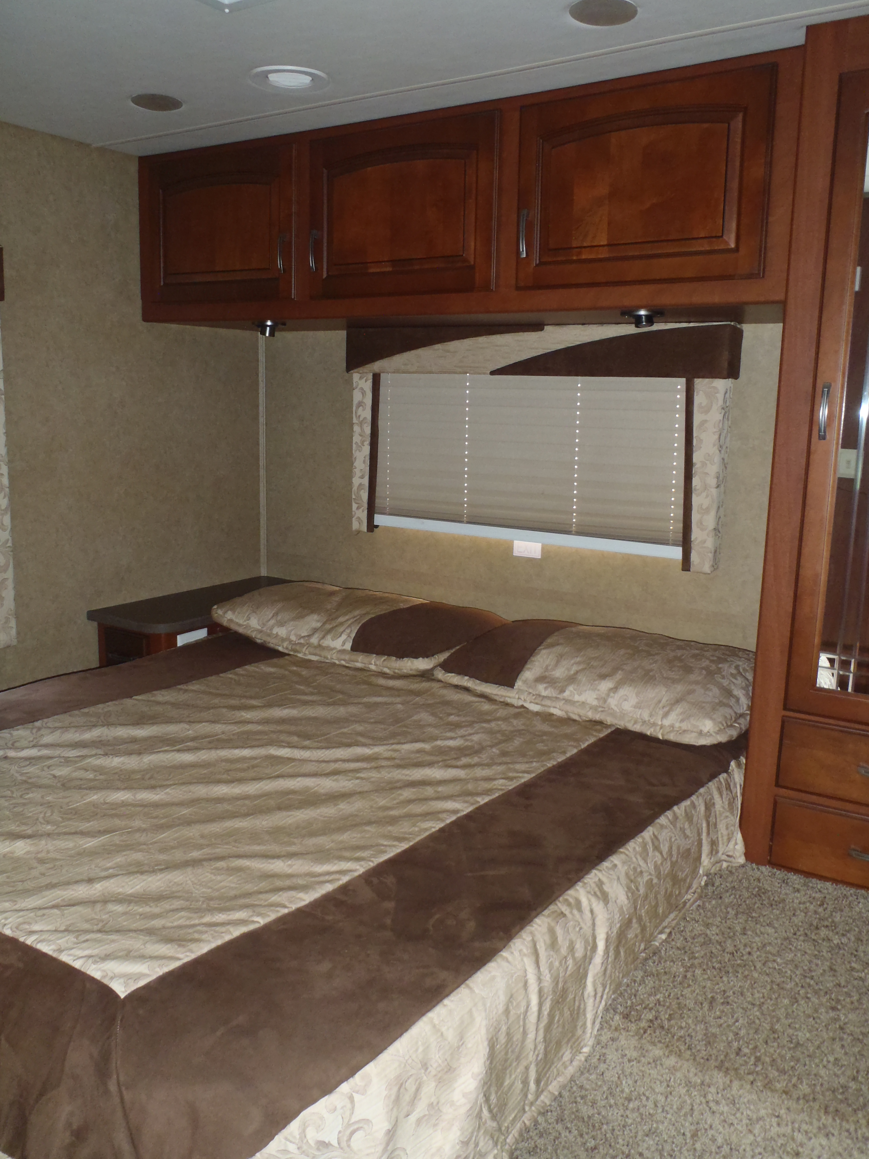 RV rental bedroom with a bed, plush brown and beige bedding, overhead wood cabinets, and a window.. Jayco GreyHawk