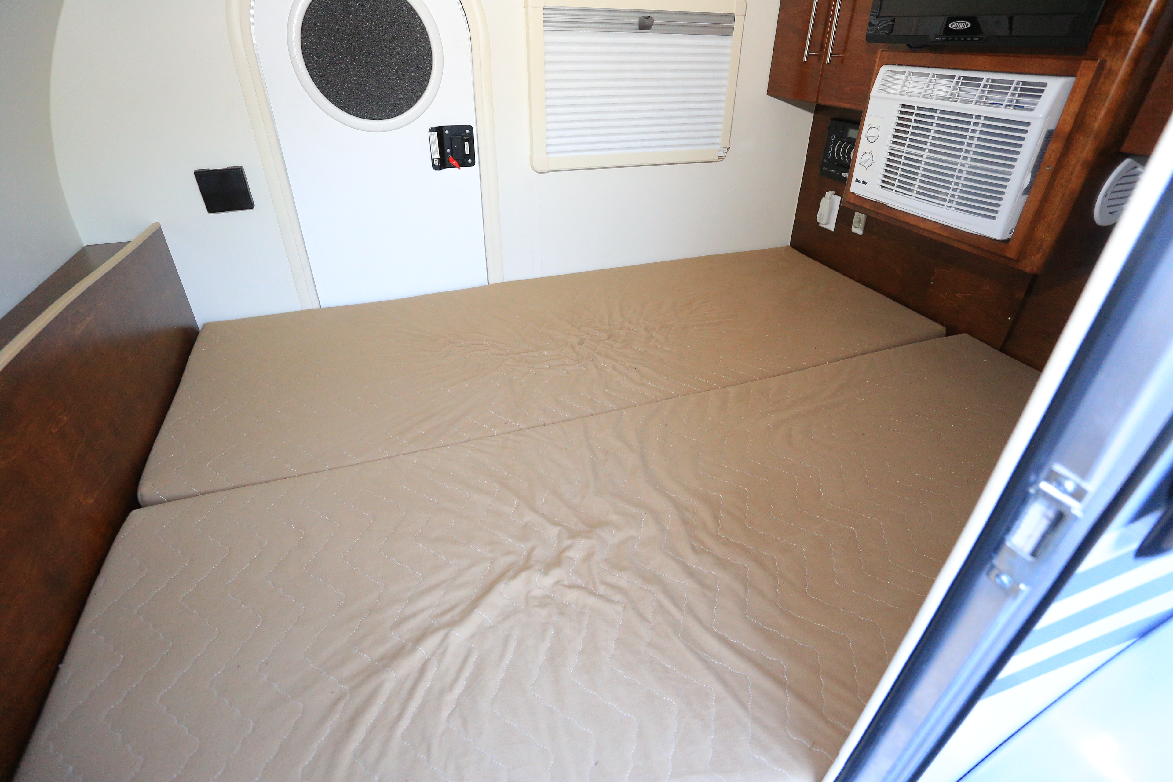 Compact rv rental interior with a light brown bed, white door with circular window, small window with blinds, and a built-in AC unit.. T@G MAX 2017