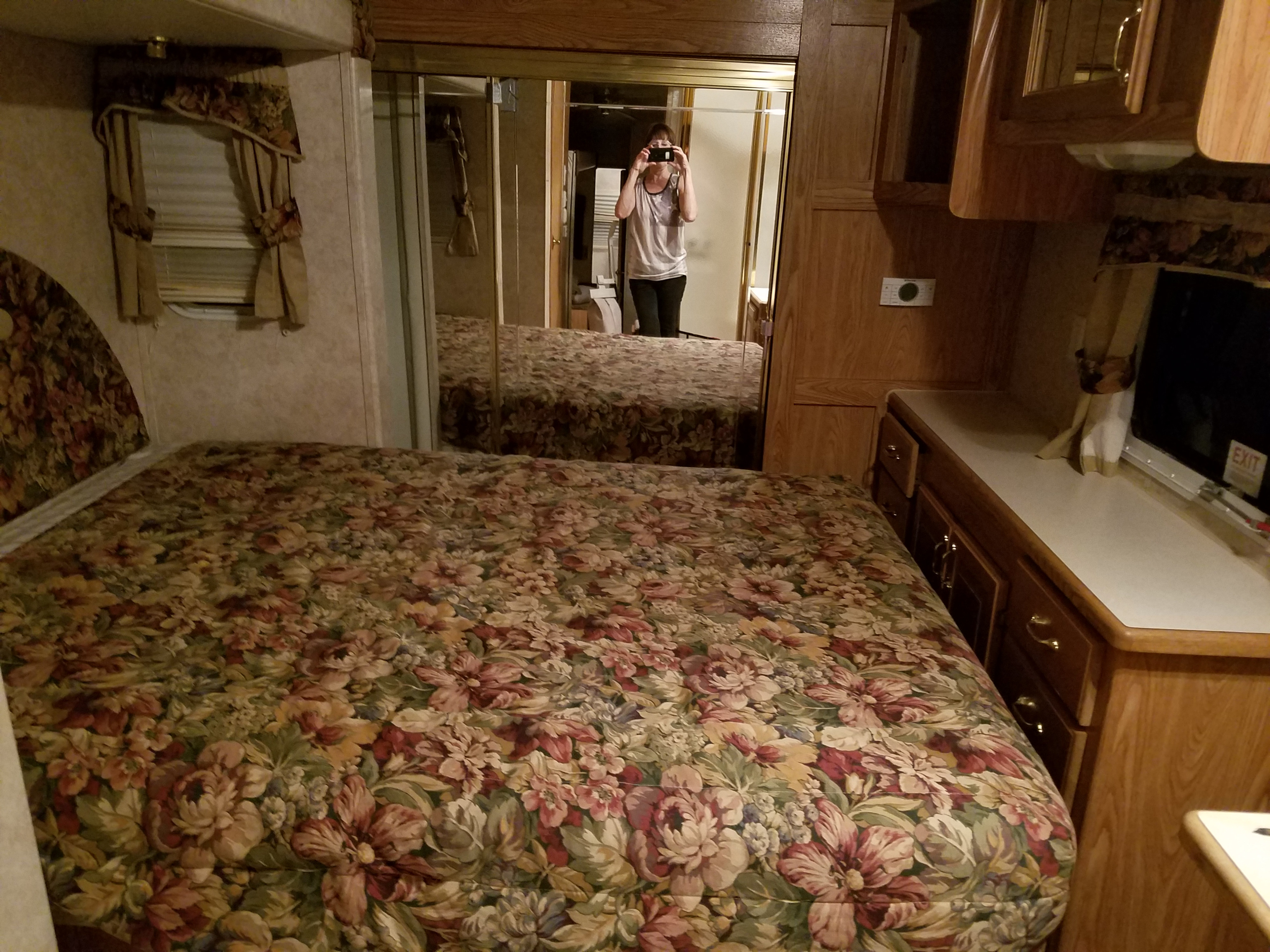 RV rental bedroom featuring a floral bed, mirrored closet, window, and wood storage. Person reflected taking photo.. Forest River Cedar Creek Silverback Edition 2005