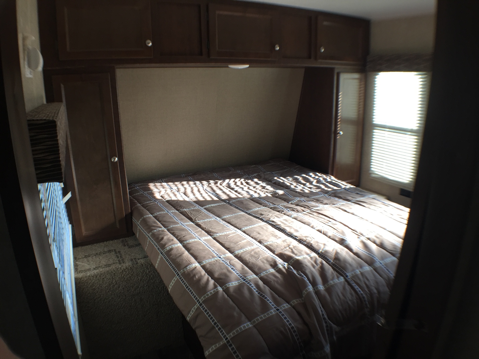 A compact RV rental bedroom with a brown bed, illuminated by sunlight stripes. Built-in cabinets are overhead.. Keystone Hideout 2017