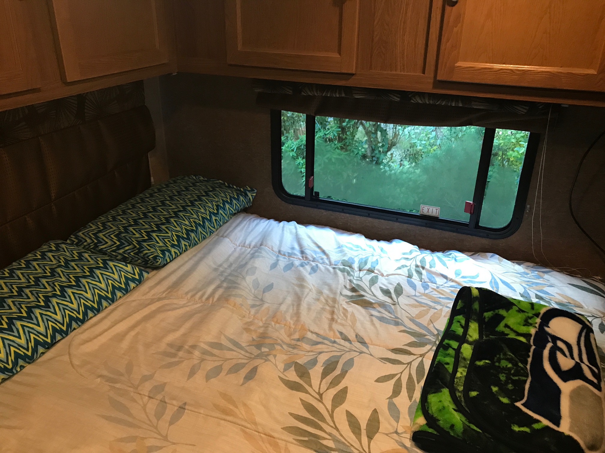 Cozy RV rental bedroom with patterned comforter and pillows. Window view shows lush greenery, with an "EXIT" sign visible.. Coachmen Leprechaun 2016