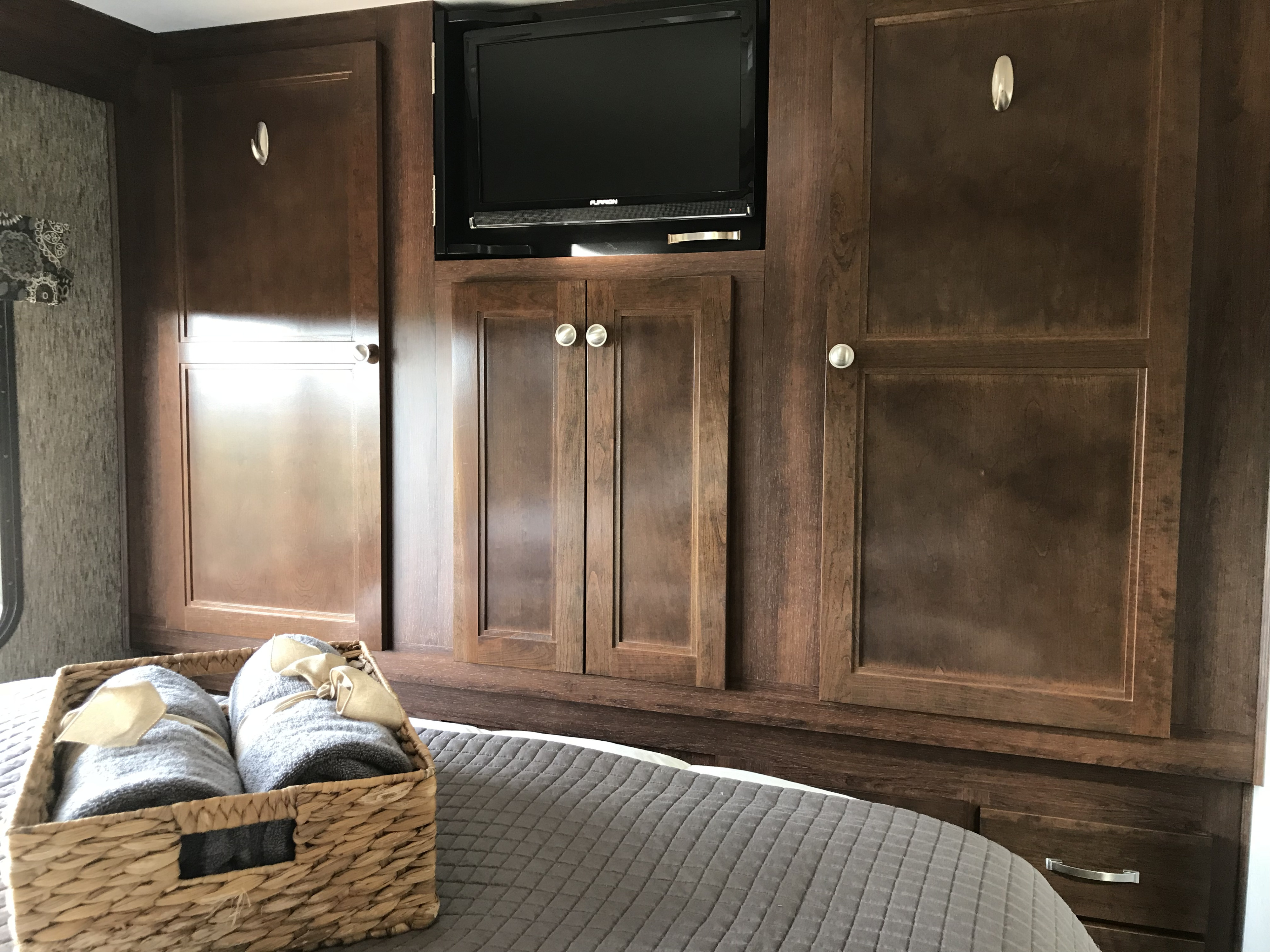 RV rental bedroom with dark wood built-in cabinetry, TV, and a bed with fresh towels in a woven basket.. Forest River Sunseeker 2018