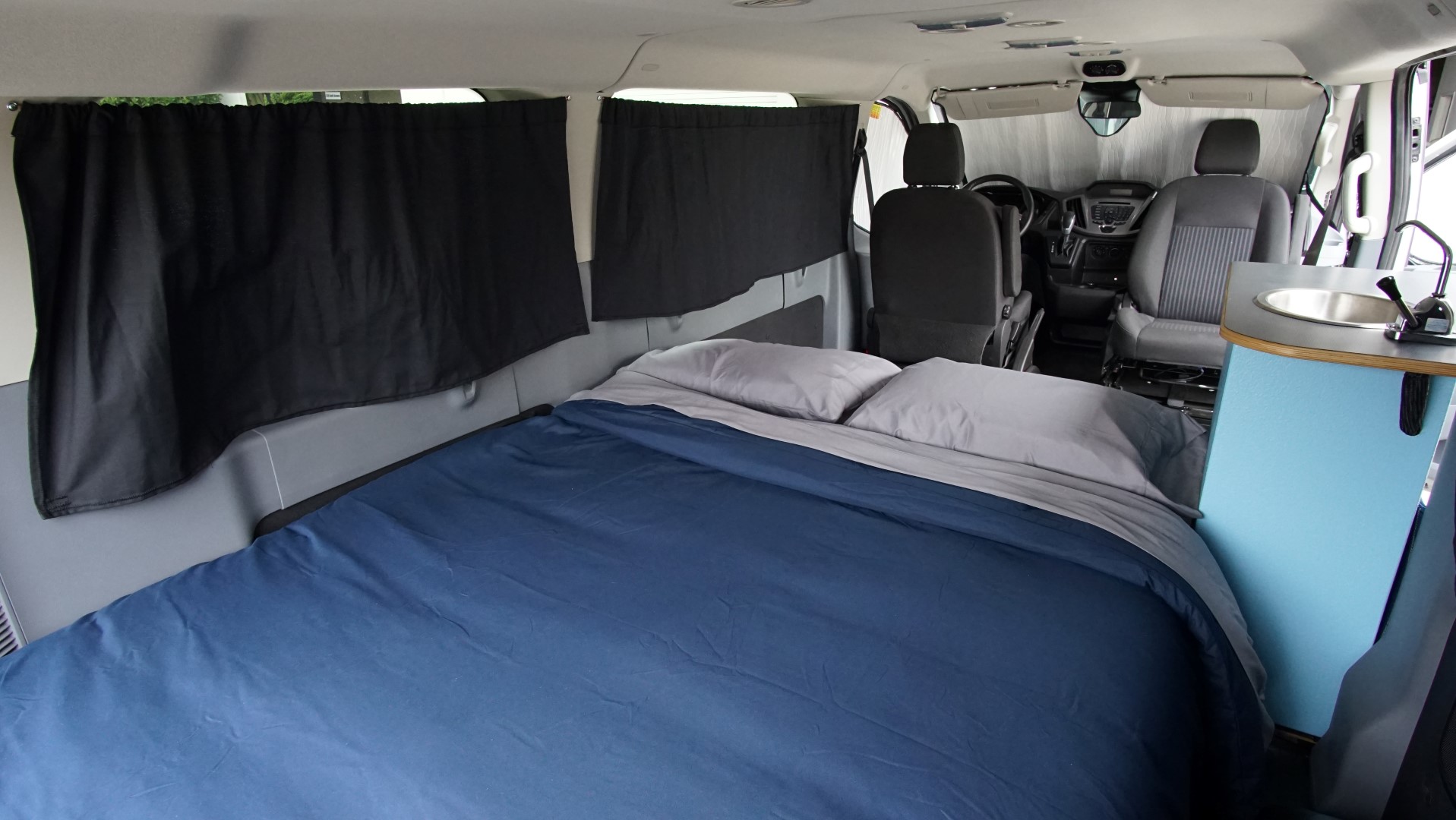 Spacious RV rental interior featuring a large bed, black privacy curtains, and a compact sink unit.. Ford Transit 2019