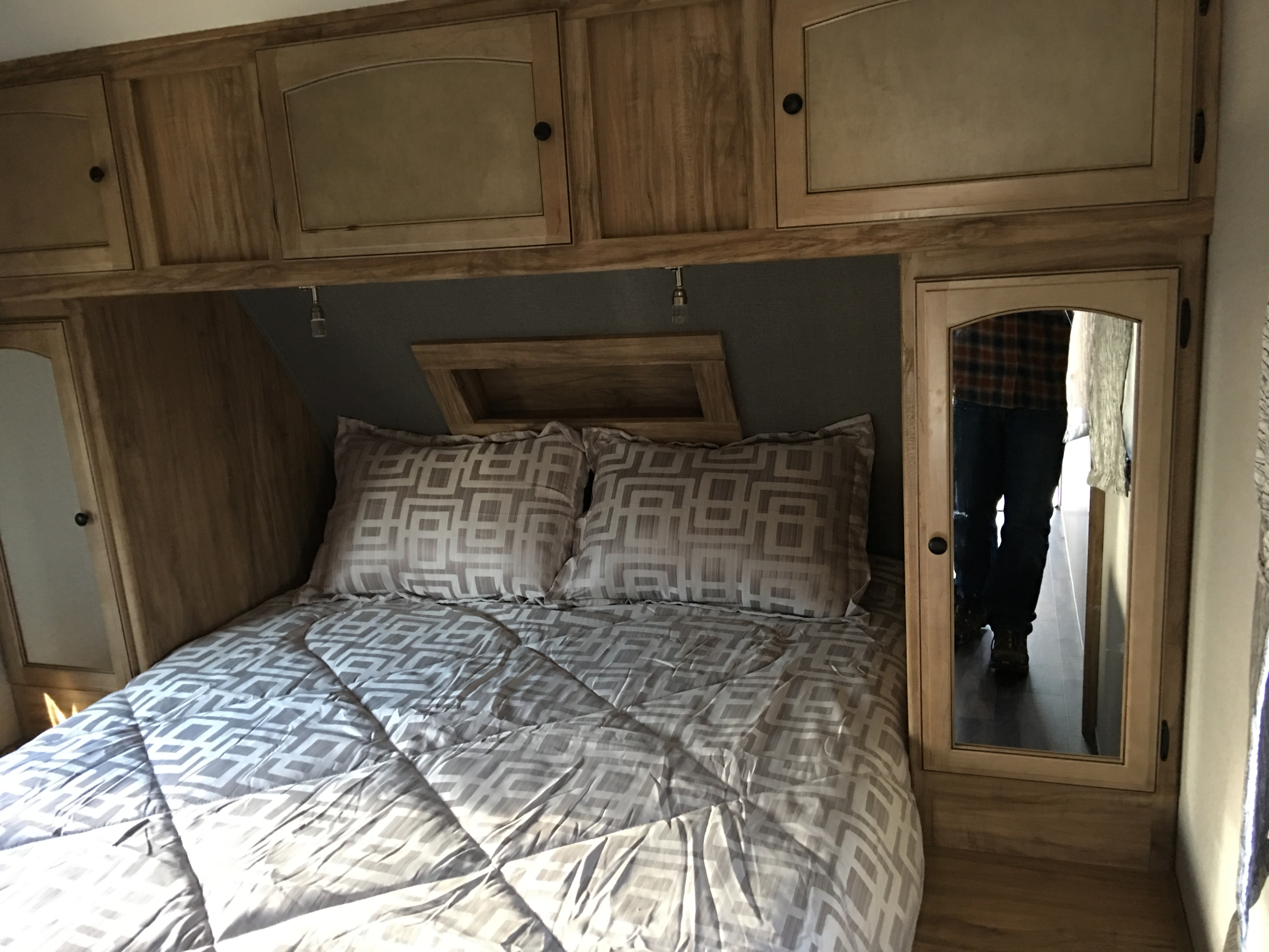 Inside an RV rental: cozy bedroom featuring a patterned bed, overhead cabinets, and a reflective mirror showing a person.. Coachmen coachman 2018