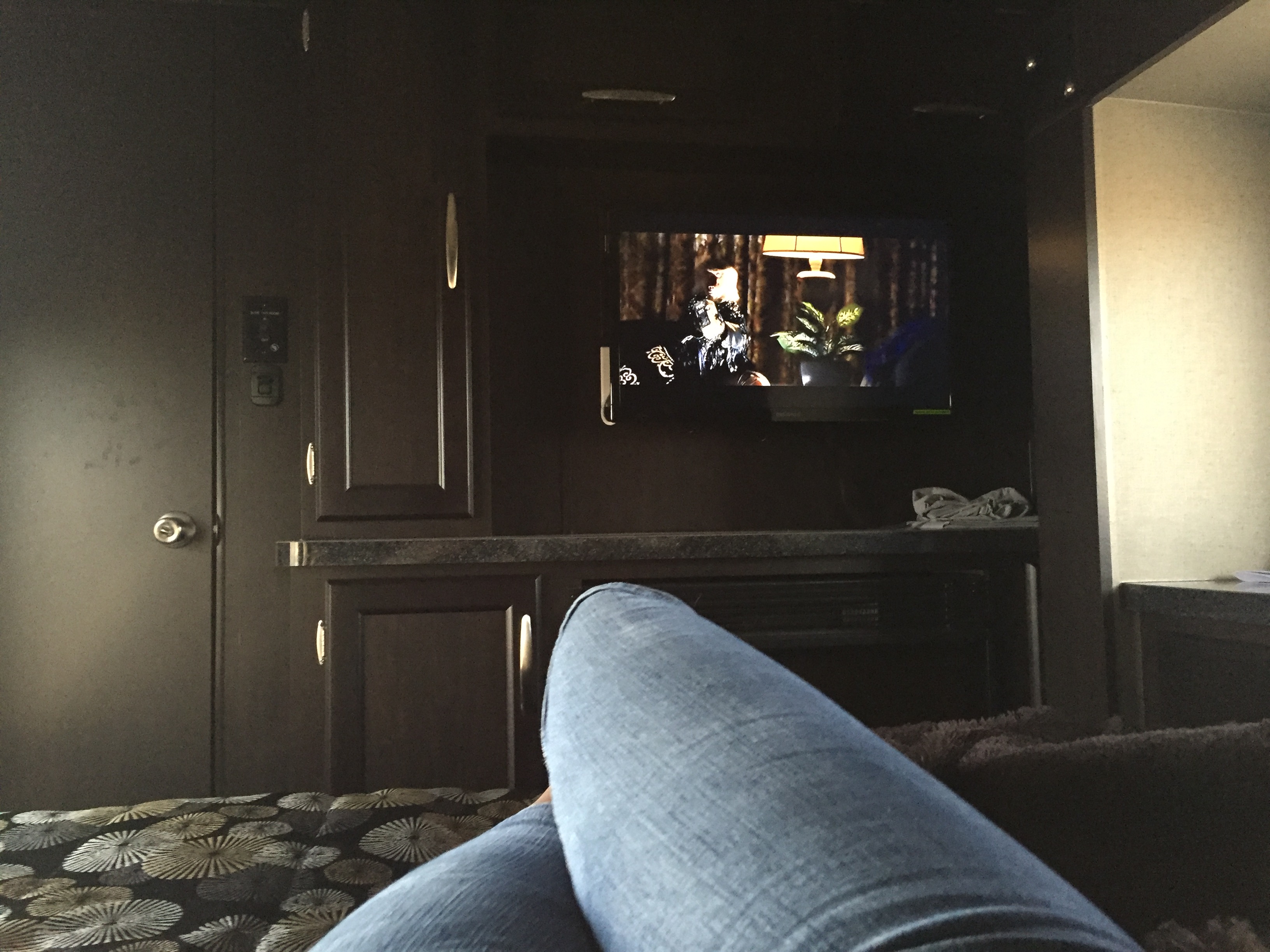Relaxing in an RV rental with jeans propped up, watching TV showing a room scene. Dark wood interior, control panel visible.. Genesis Supreme Rv 37GS Toy Hauler 2017
