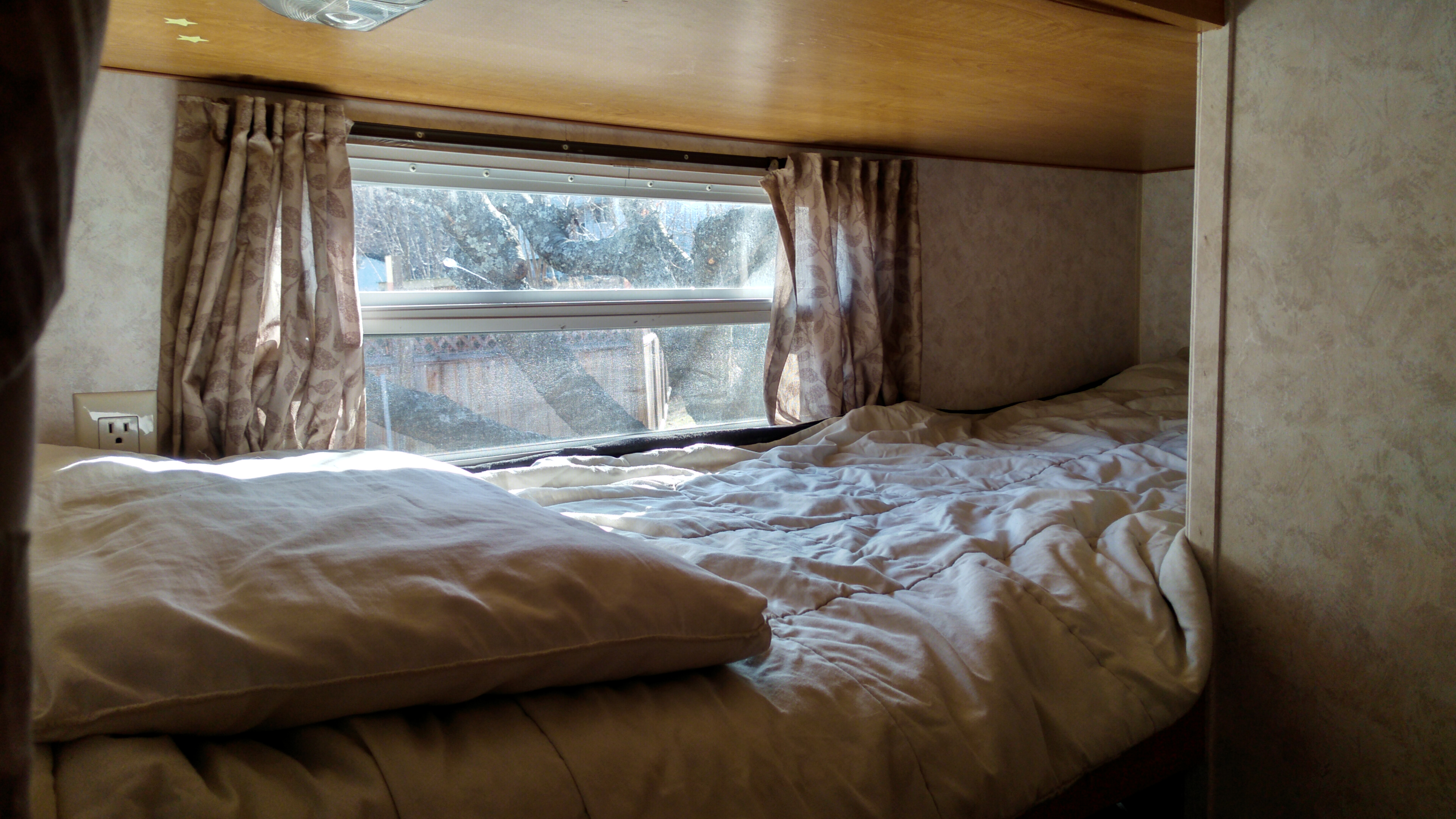 Cozy RV rental bunk bedroom with a sunlit window, bed, and outlet, providing a comfortable, compact sleeping area.. Layton Layton Trailer 2007