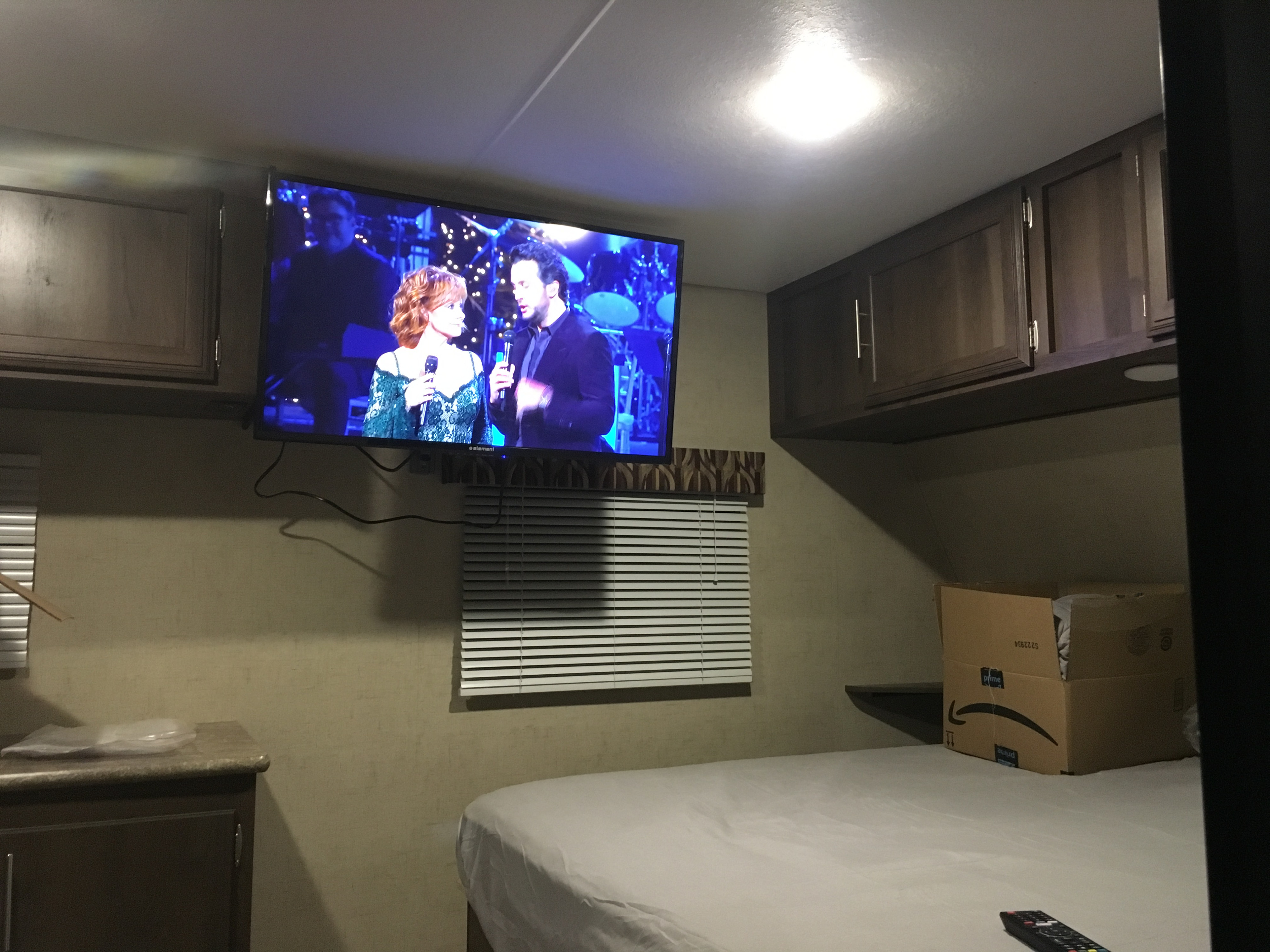 An RV rental interior features a TV with performers, a comfy bed, wood cabinets, and window blinds.. KZ SPORTSMEN 2017