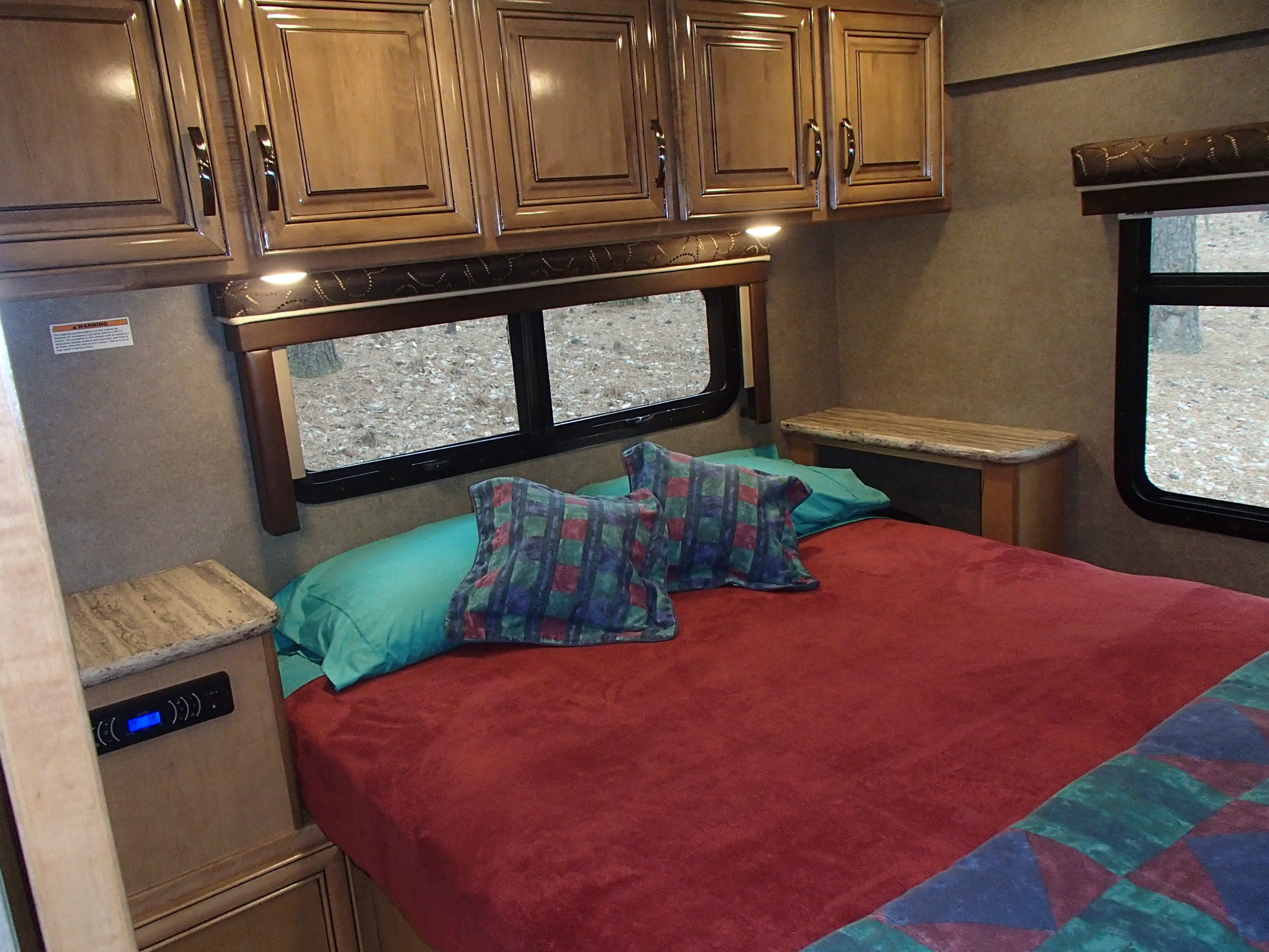Cozy RV rental bedroom with a red bed, teal pillow, wooden cabinets, and outdoor views from the windows.. Thor Quantum 2017
