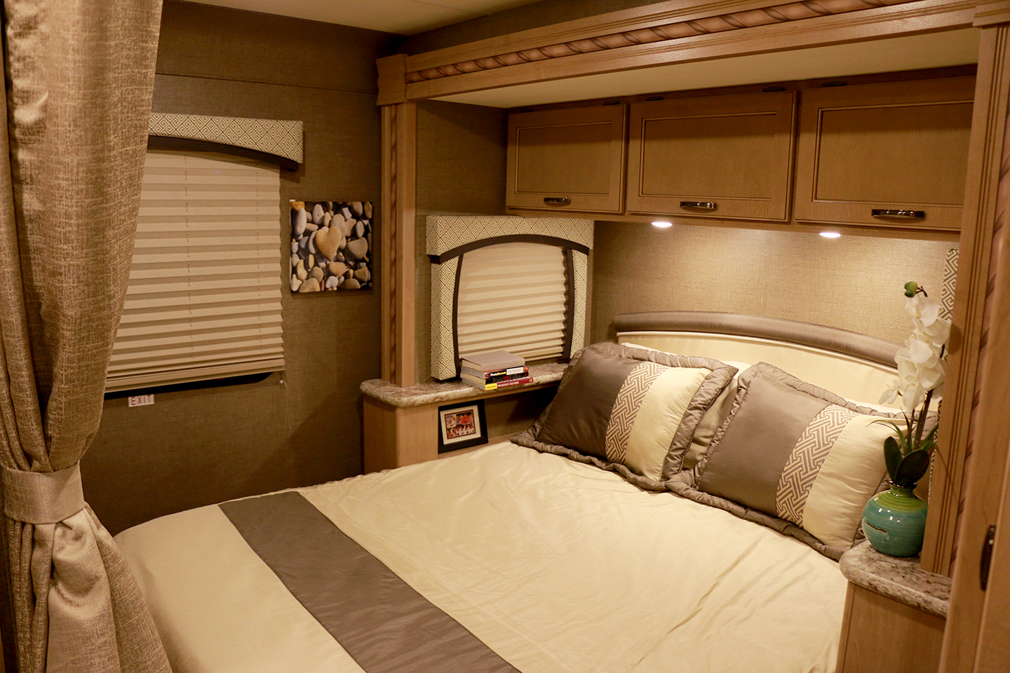 This comfortable RV rental bedroom features a plush bed, overhead cabinets, windows with blinds, and warm lighting.. Thor Motor Coach Chateau 2018